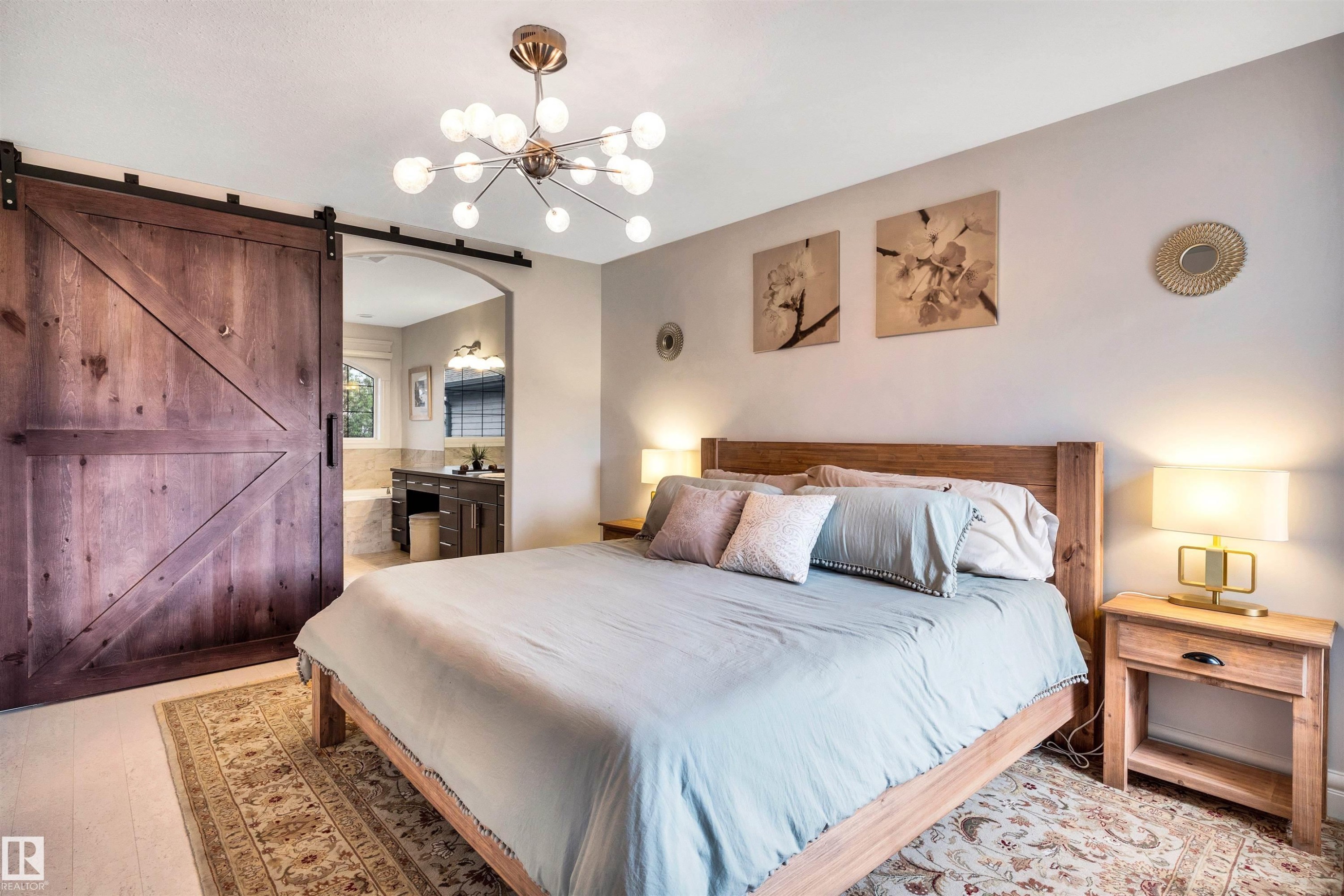 Spacious sleeping quarters featuring a modern ceiling light fixture, light wood-finish flooring, and a large barn door providing access to a bathroom with a built-in vanity and soaking tub - 11935 21 Avenue, Edmonton, AB