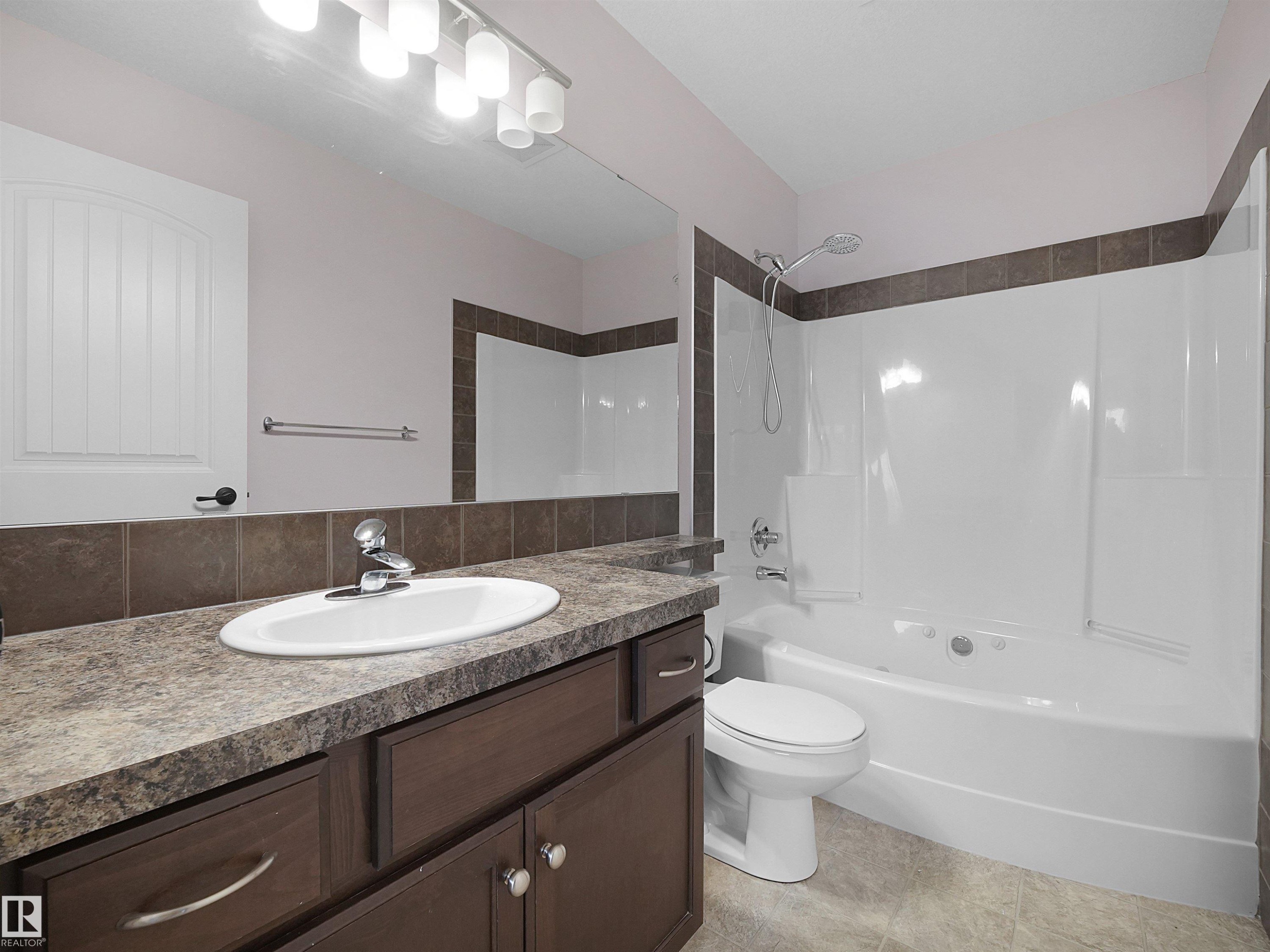 Bathroom featuring a white oval sink set into a speckled countertop, dark wood-finish cabinetry, a white bathtub with a built-in shower, brown tile accents, and a white toilet - 21316 45A Avenue Nw, Edmonton, AB