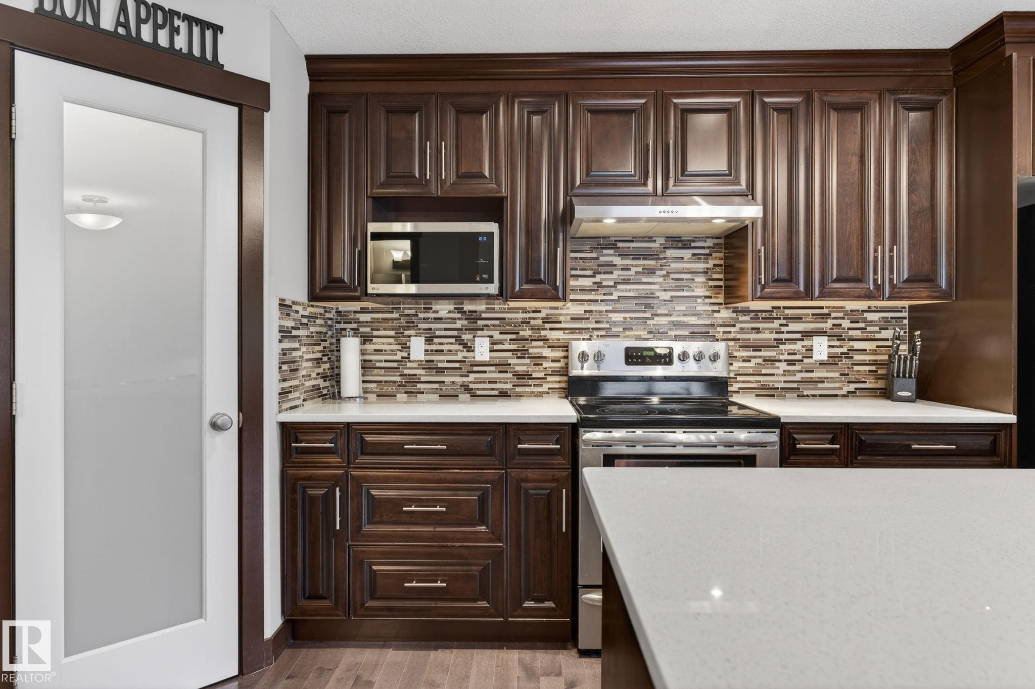 Kitchen featuring dark wood cabinetry with crown molding, stainless steel appliances, a mosaic tile backsplash, white countertops, and wood-finish flooring - 8506 80 Avenue, Edmonton, AB