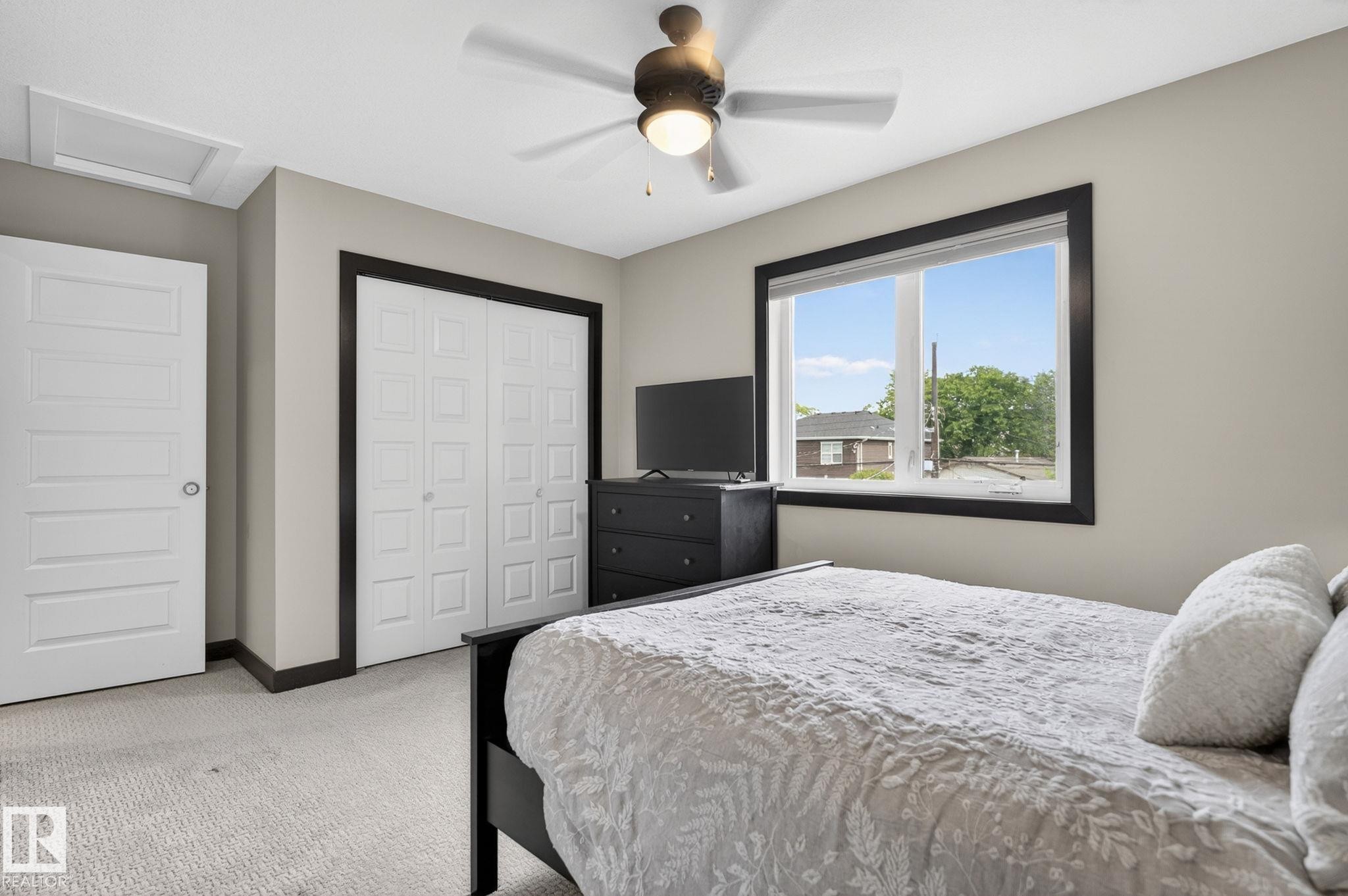 Neutral-toned room featuring a ceiling fan with integrated lighting, a broad window with dark trim, light-toned carpet, and a white paneled door - 8506 80 Avenue, Edmonton, AB