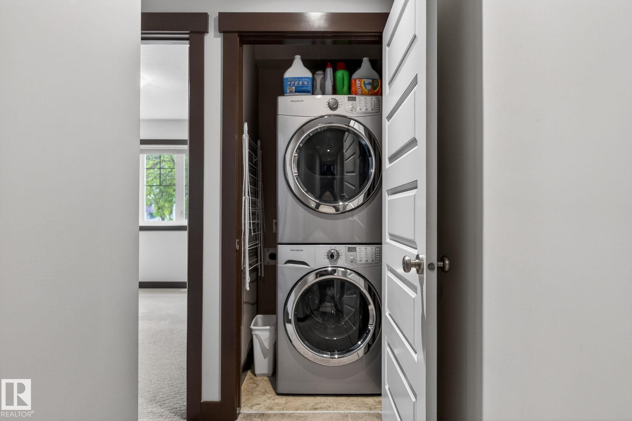 Stacked laundry appliances within a dedicated closet, featuring a white louvered door, dark wood trim, and tile flooring - 8506 80 Avenue, Edmonton, AB