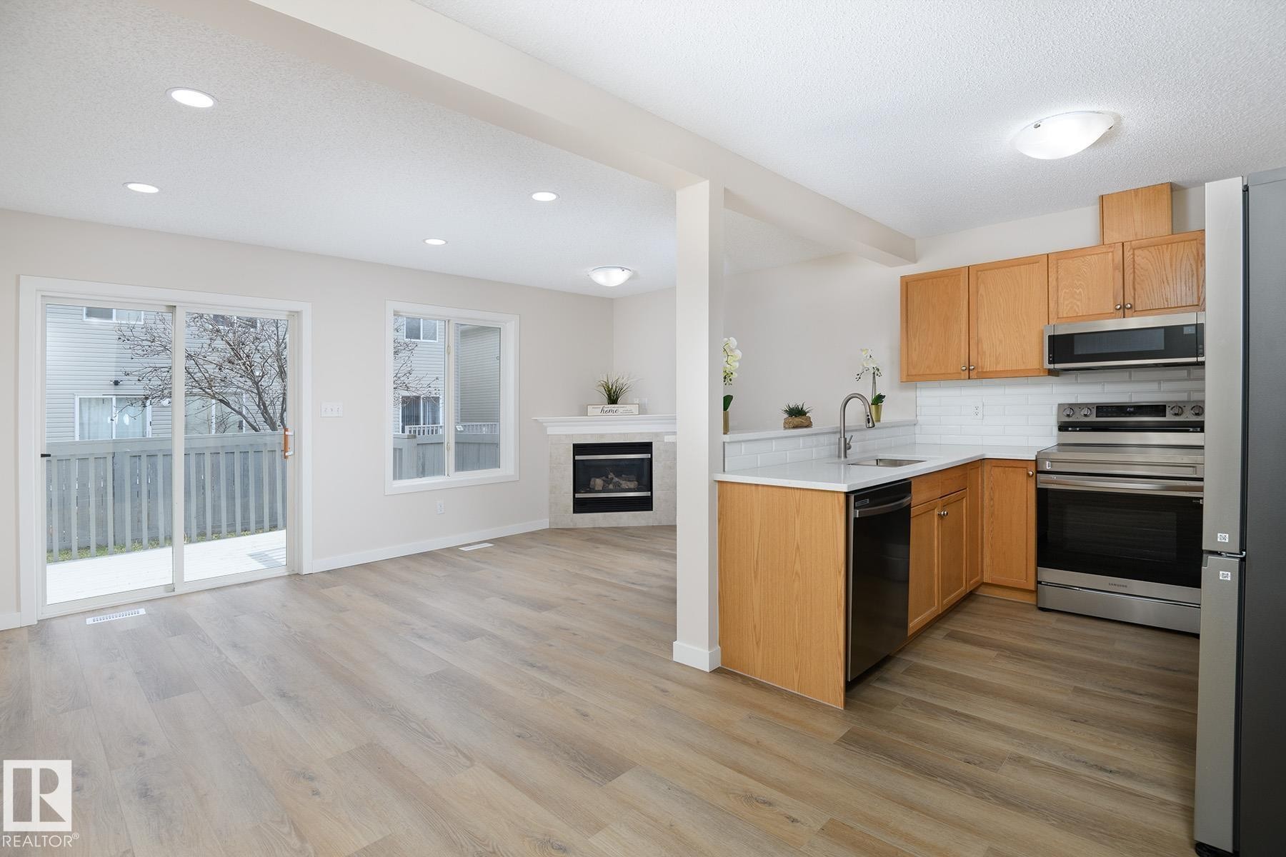 Open-concept kitchen and living area featuring wood-finish flooring, a white subway tile backsplash, stainless steel appliances, a built-in fireplace, and sliding glass doors to a private deck - #55 14603 Miller Bv Nw, Edmonton, AB