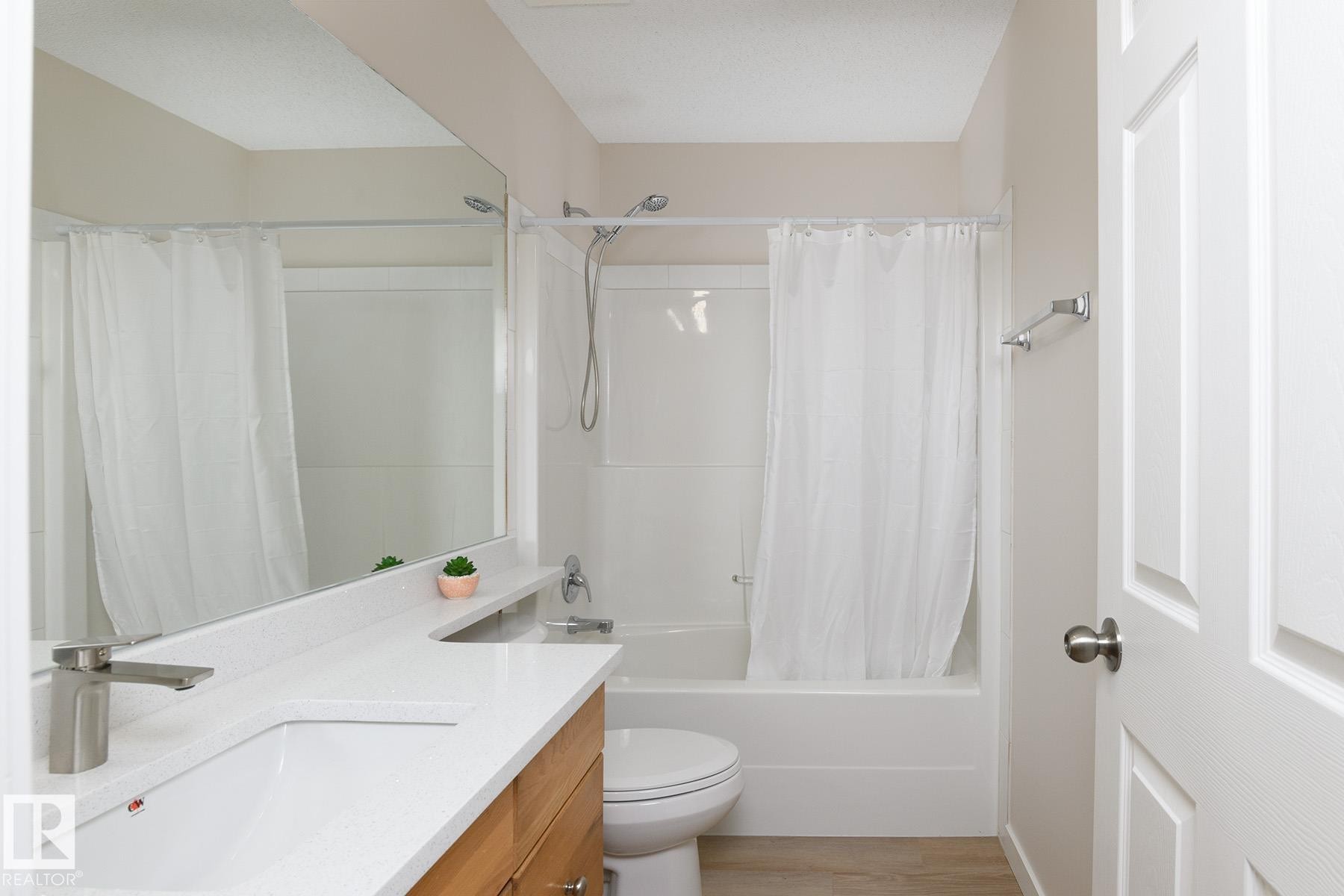White tub-shower combination with a single-handle faucet, integrated white countertop with a rectangular sink, wood-finish vanity cabinetry, and a large wall-mounted mirror - #55 14603 Miller Bv Nw, Edmonton, AB