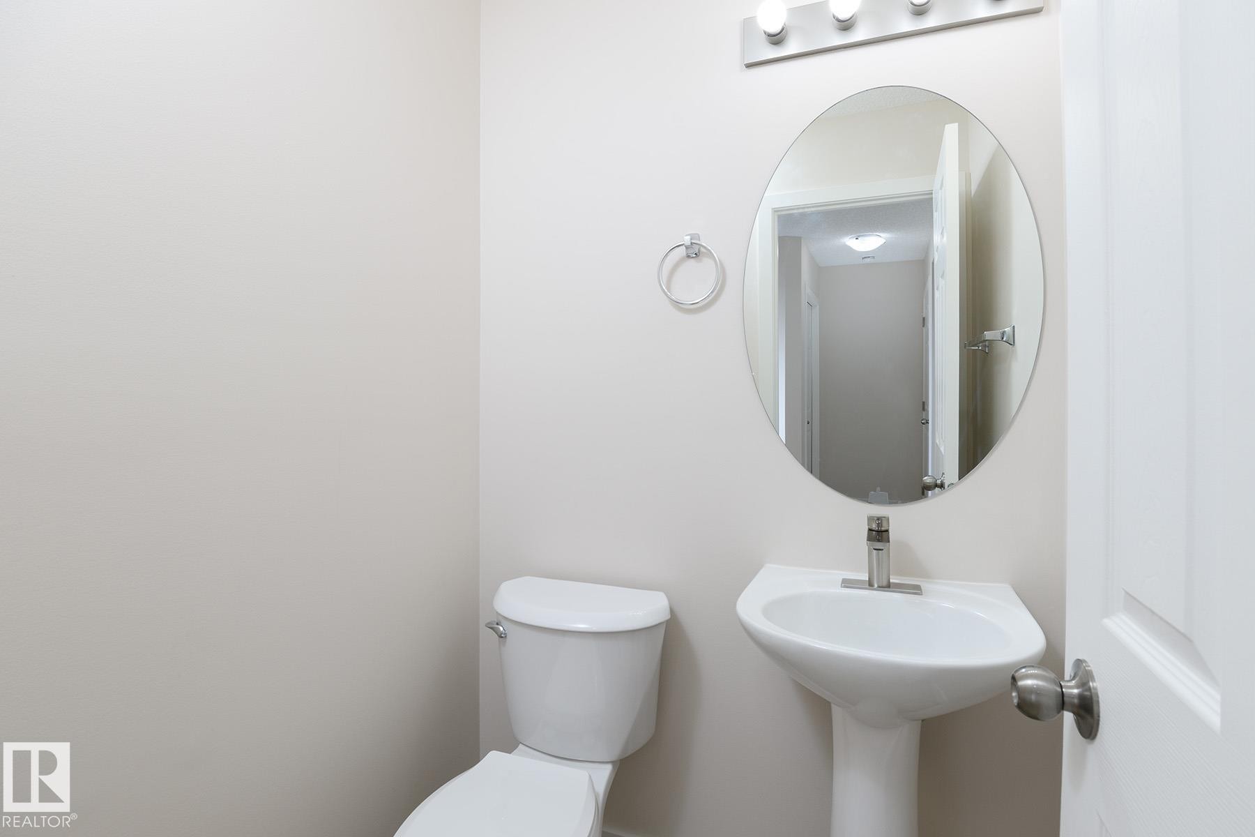 Powder room featuring a pedestal sink with a single-handle faucet, oval mirror, and a three-light vanity fixture - #55 14603 Miller Bv Nw, Edmonton, AB