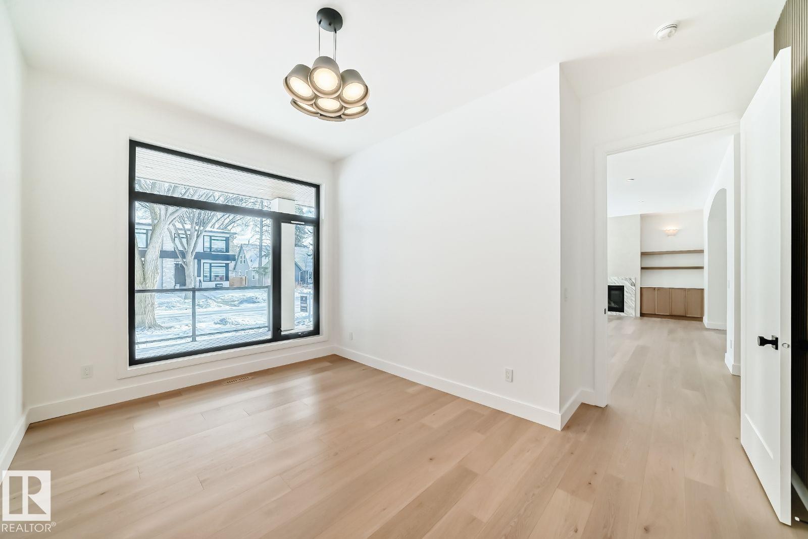 Spacious room with light wood-finish flooring, a contemporary chandelier, a large black-framed window, bright white walls, and a wide entryway - 10514 134 Street, Edmonton, AB