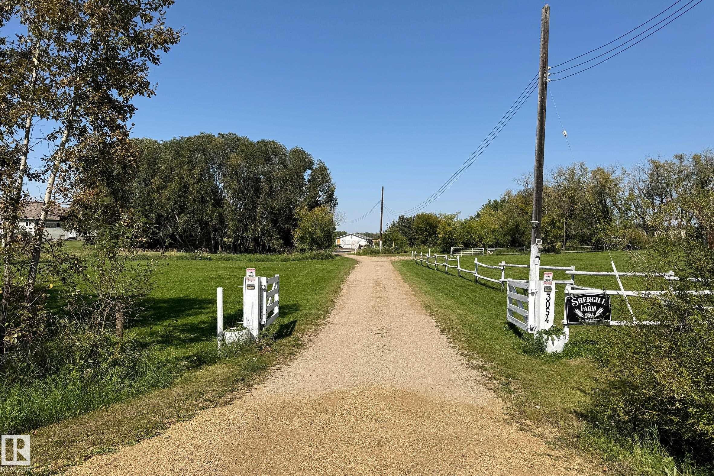 Gravel driveway leading to a distant structure, bordered by green lawns and a white split-rail fence - 3054 28 Avenue, Edmonton, AB