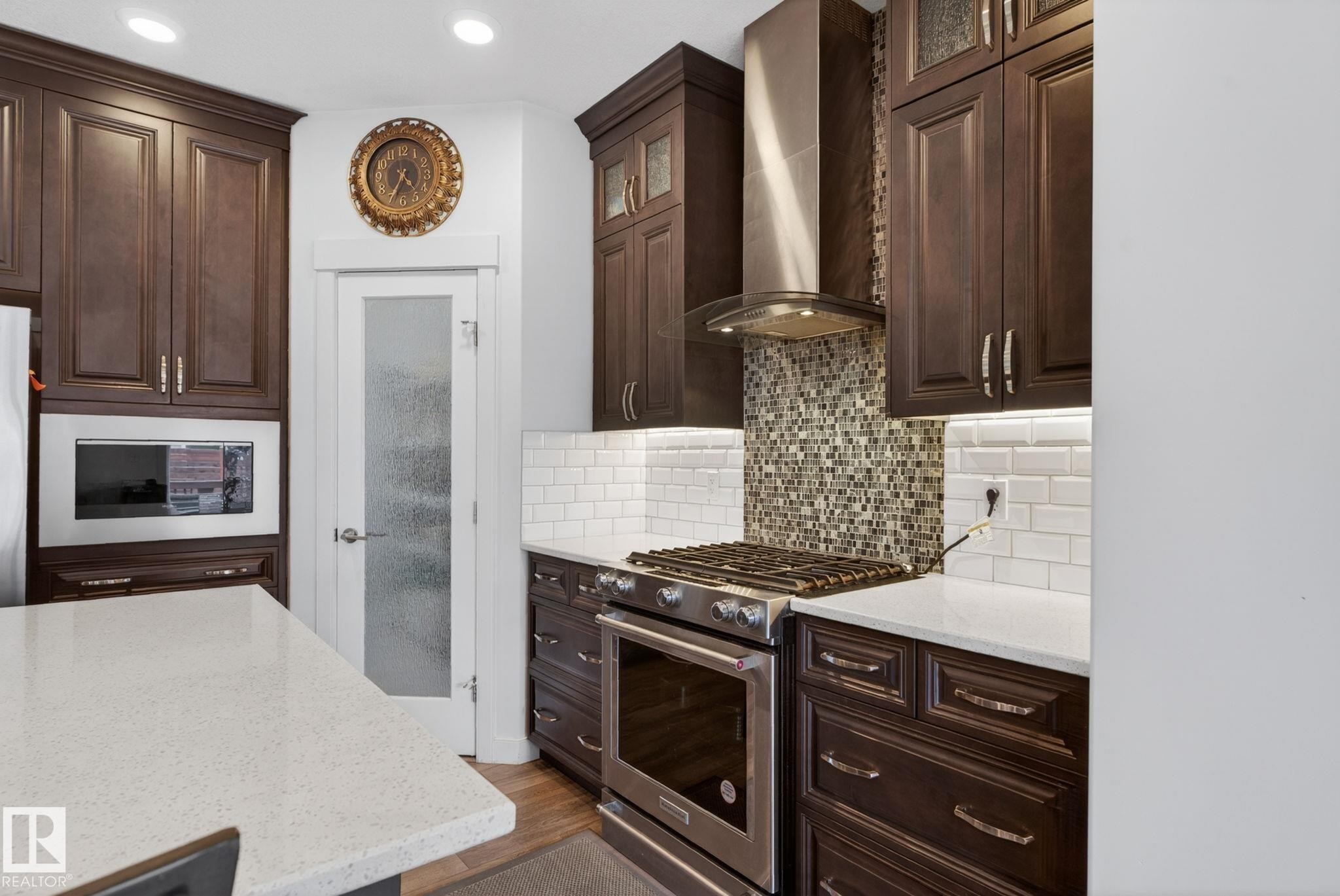 Kitchen featuring dark wood cabinetry, white quartz countertops, subway tile backsplash, stainless steel appliances, and a frosted glass pantry door - 704 39 Street, Edmonton, AB