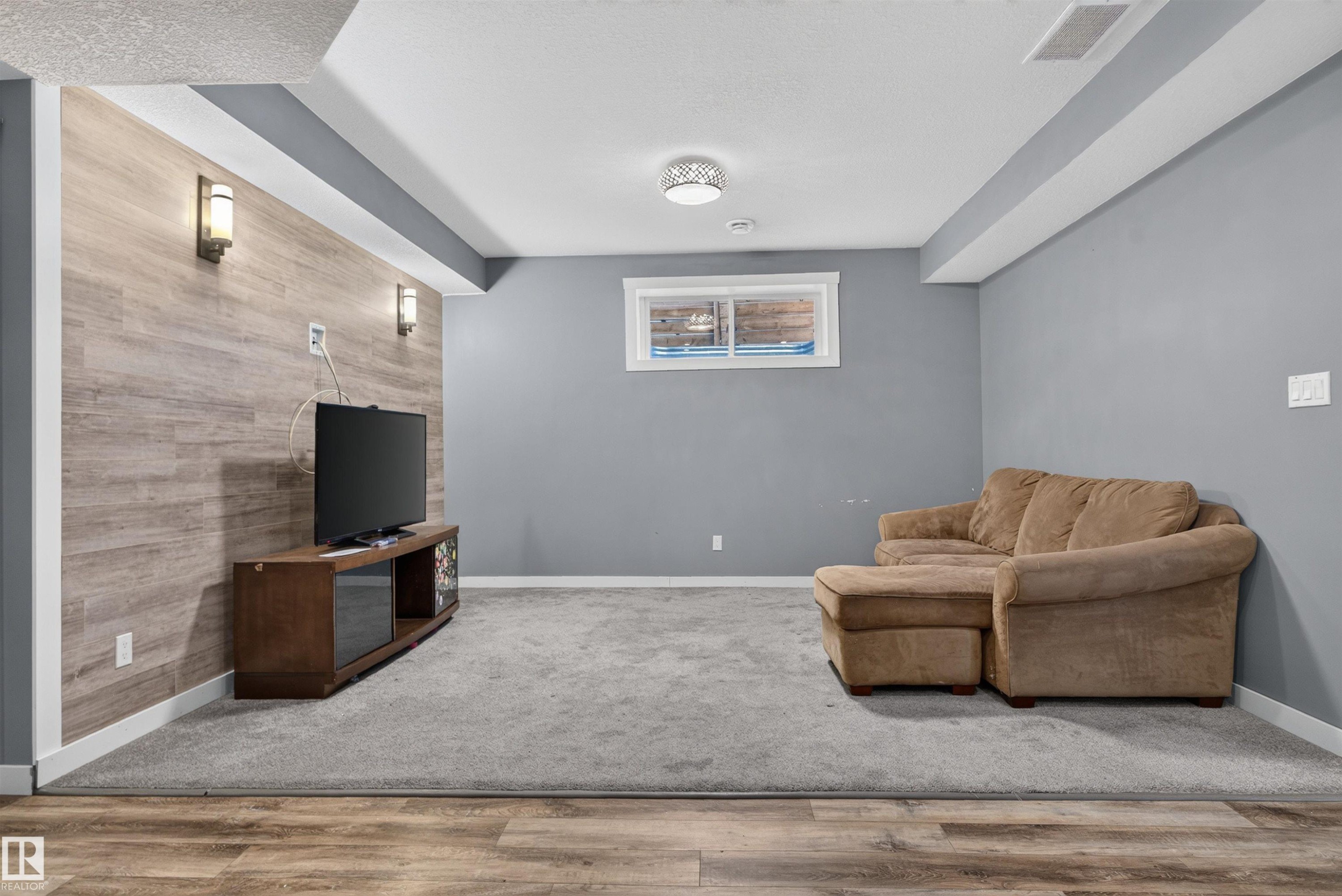 Finished room featuring wood-finish accent wall with sconce lighting, neutral carpet flooring, a white-framed window, and overhead lighting - 704 39 Street, Edmonton, AB