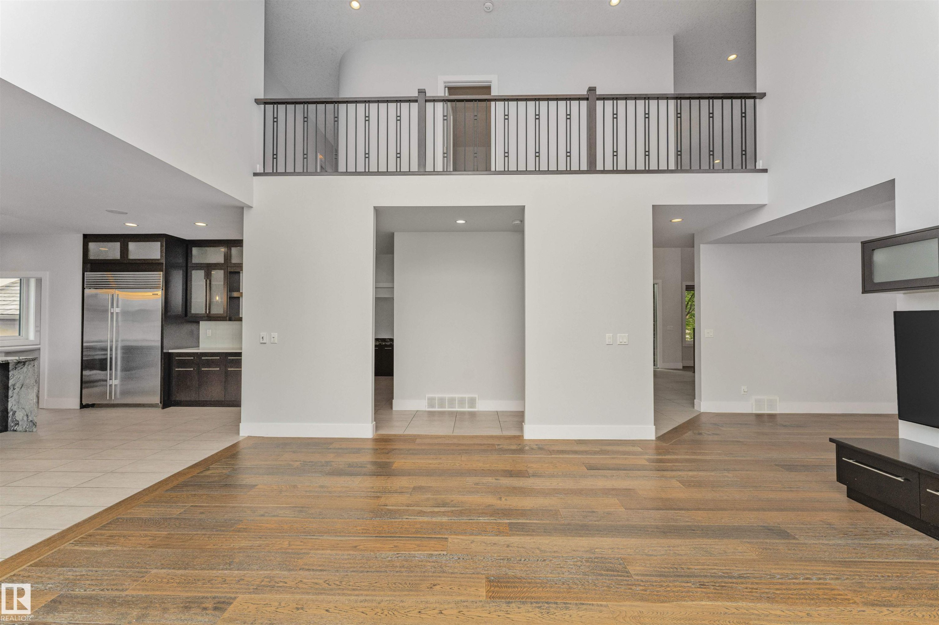 Open-concept living space featuring wood-finish flooring, a second-story interior balcony with wrought iron railing, recessed lighting, and a partial view of a kitchen with dark cabinetry and stainless steel appliances - 3310 Watson Bay, Edmonton, AB