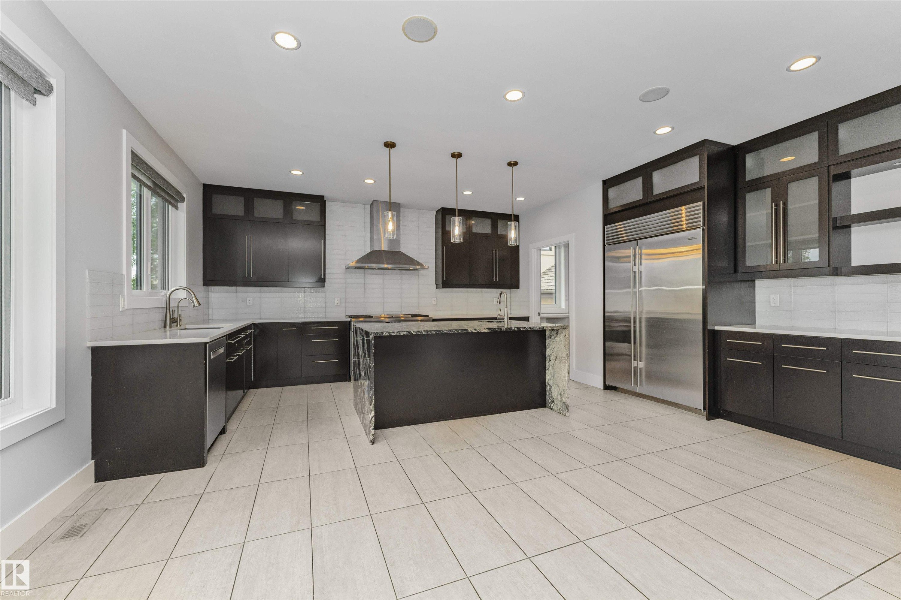 Open-concept kitchen featuring dark wood cabinetry, stainless steel appliances, a center island with a waterfall countertop, recessed lighting, and large format tile flooring - 3310 Watson Bay, Edmonton, AB