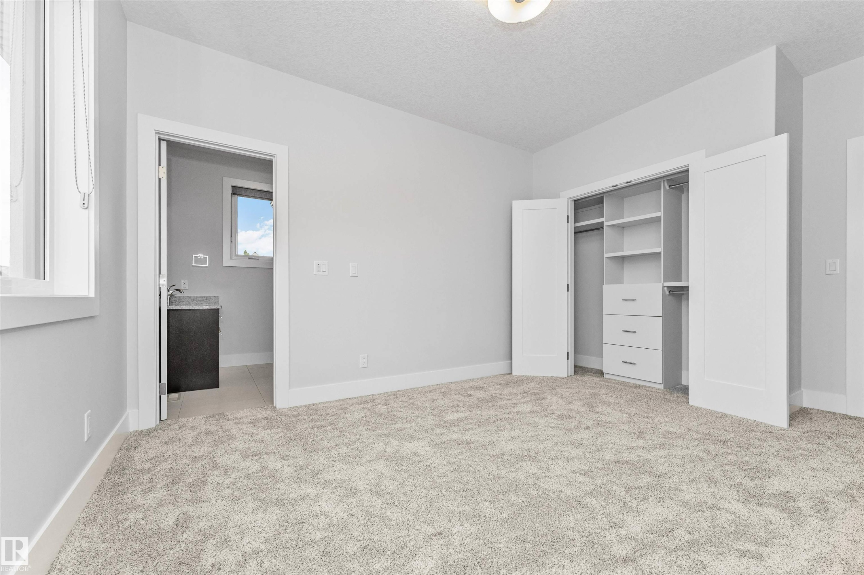 Spacious room featuring light gray walls, plush carpeting, a built-in closet system with shelving and drawers, a window with white trim, and a doorway revealing a vanity with a dark countertop and a small window - 3310 Watson Bay, Edmonton, AB