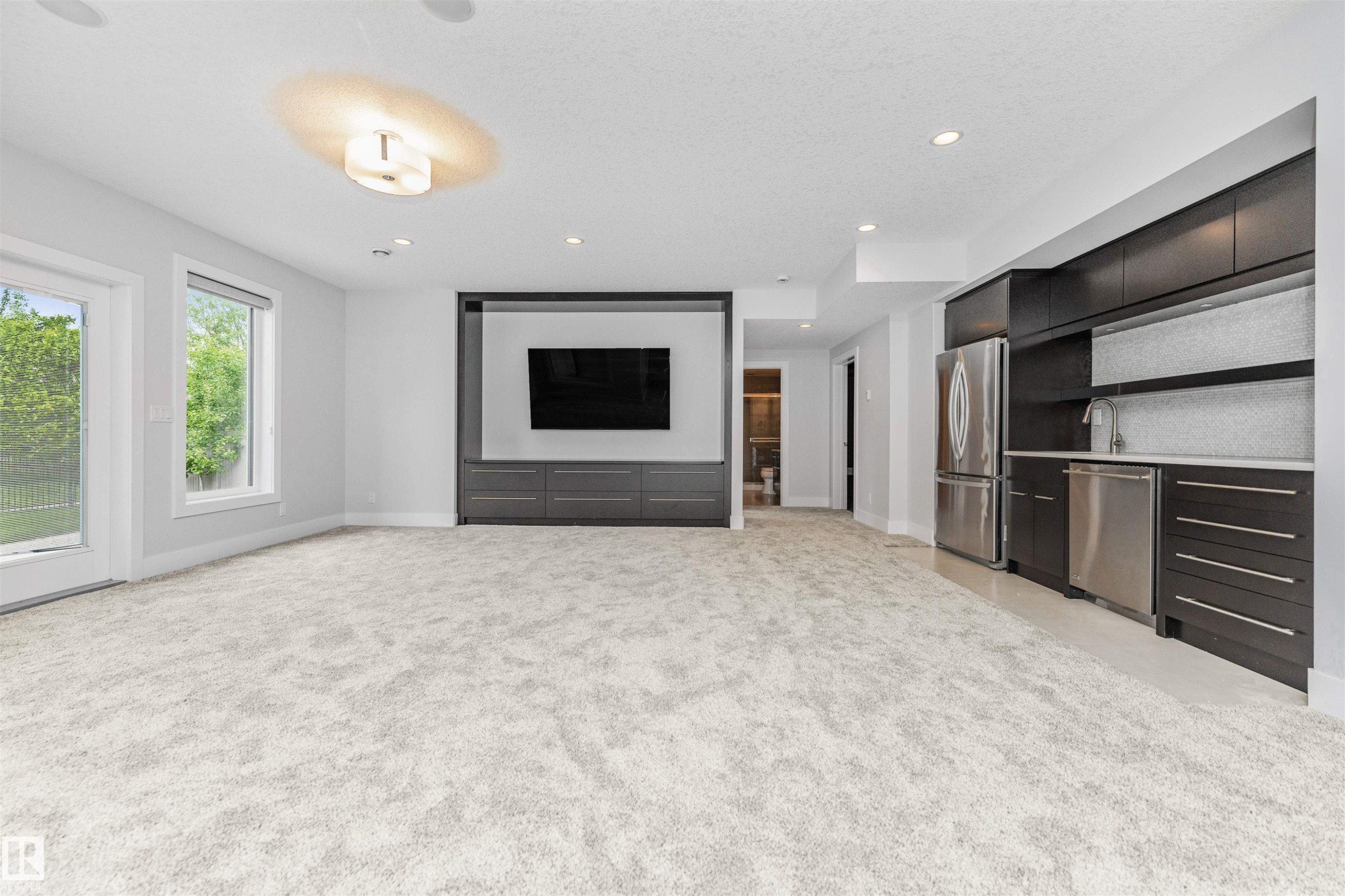 Spacious entertainment area featuring light-toned carpeting, a built-in media center with integrated cabinetry, and a wet bar complete with stainless steel appliances, dark wood-finish cabinetry, and a light-toned countertop - 3310 Watson Bay, Edmonton, AB