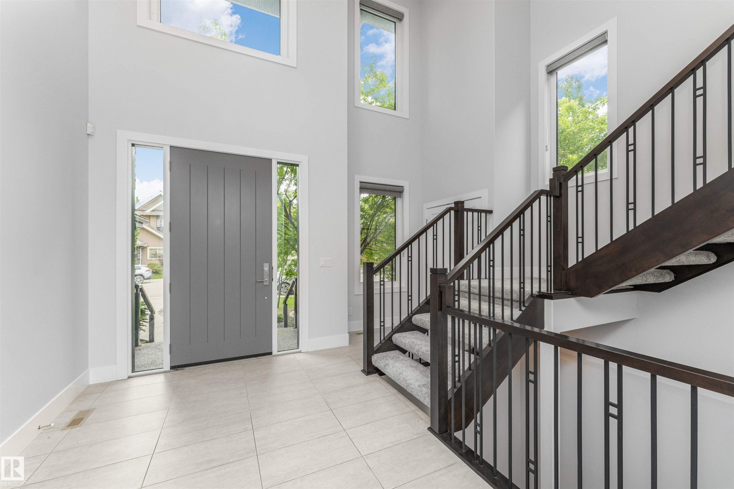 Two-story foyer featuring a gray panel door with sidelights, a custom dark wood-finish staircase with wrought iron railings, and light-toned tile flooring - 3310 Watson Bay, Edmonton, AB
