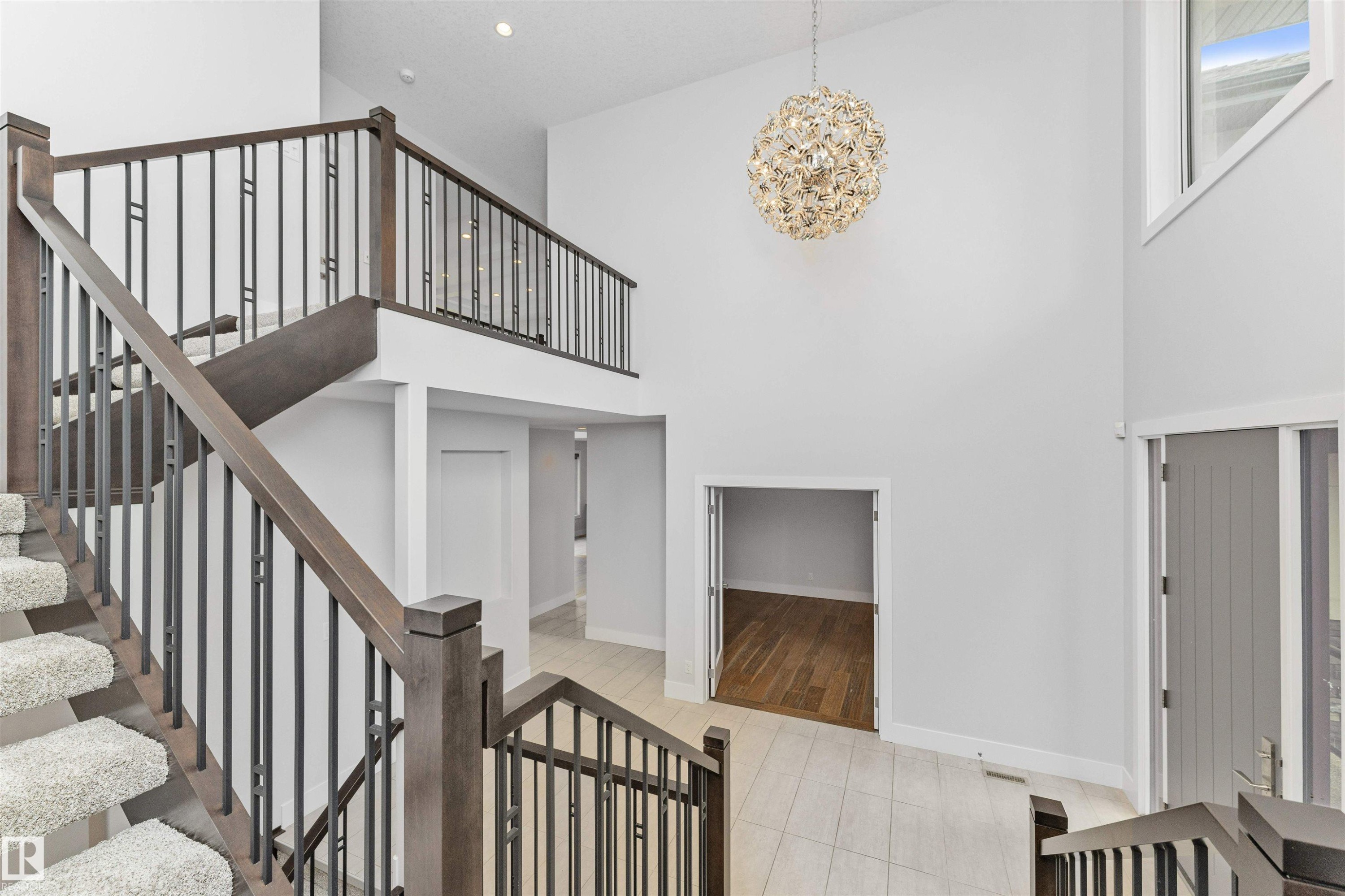 Two-story foyer featuring a contemporary chandelier, a multi-level staircase with wood-finish handrails and black metal balusters, and light-toned tile flooring - 3310 Watson Bay, Edmonton, AB
