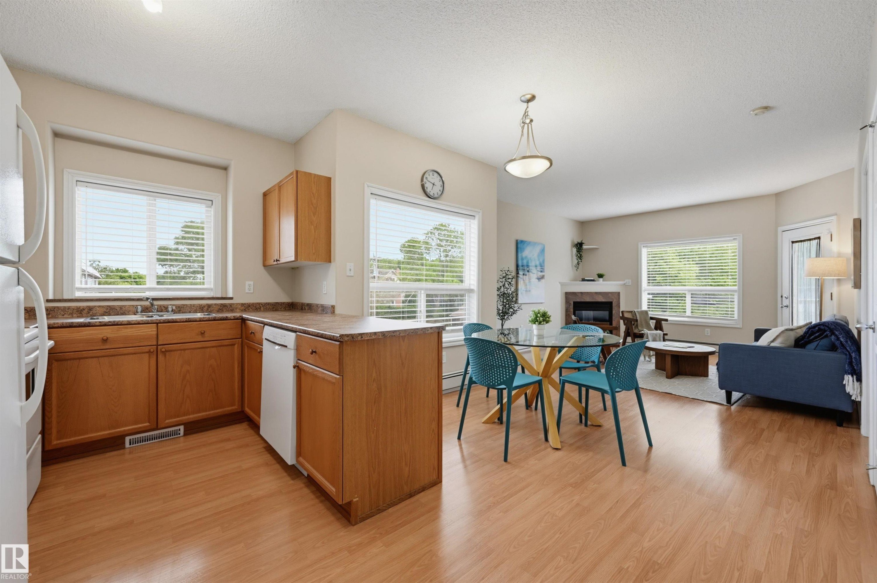 Open-concept living area featuring wood-finish flooring, a kitchen with light wood cabinetry and a peninsula, a dining area with pendant lighting, a living room with a fireplace, and multiple windows with blinds - 203 10719 80 Avenue, Edmonton, AB