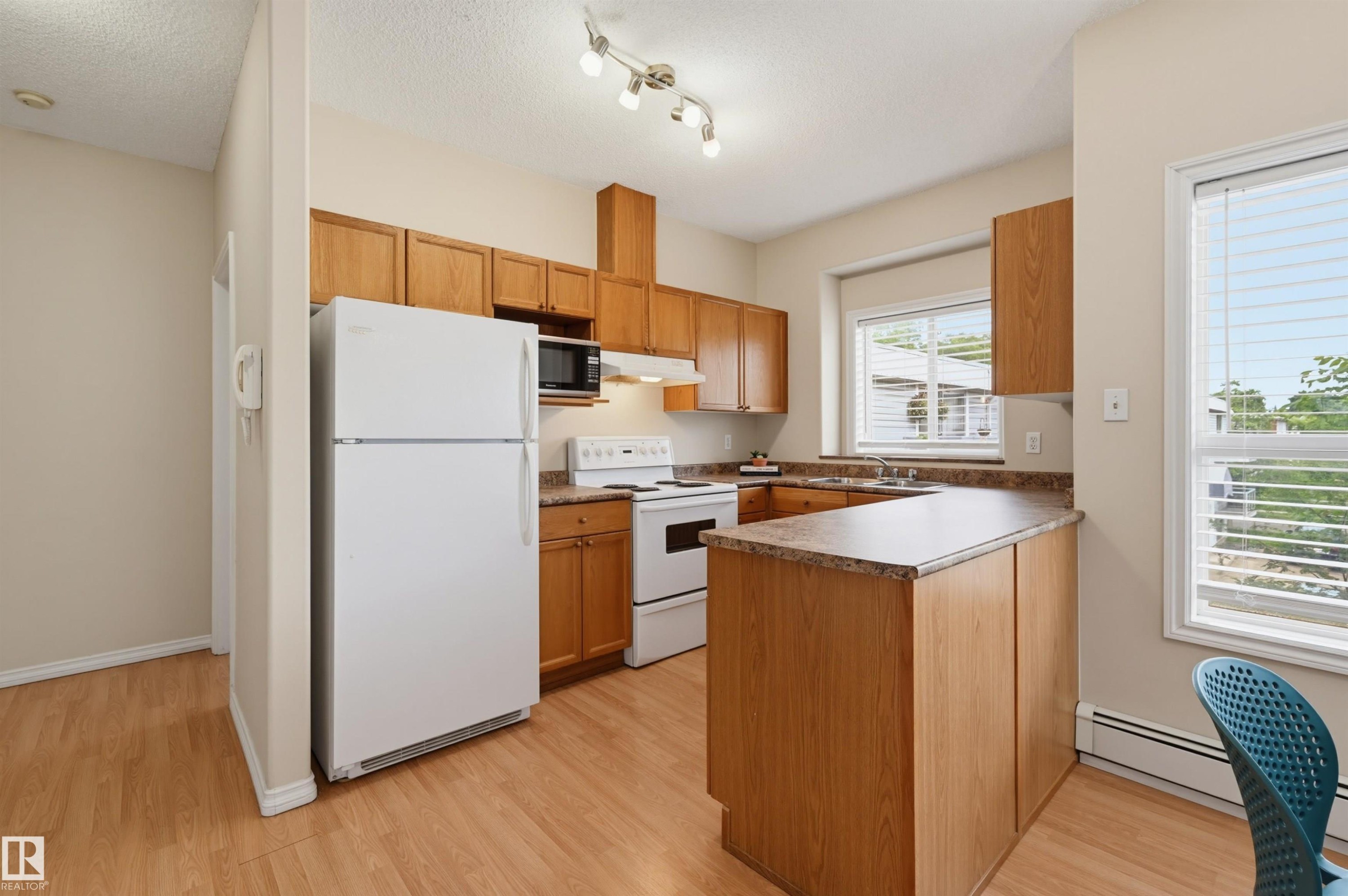 Kitchen featuring wood-finish flooring, wood cabinetry, white appliances, track lighting, and a window with blinds - 203 10719 80 Avenue, Edmonton, AB