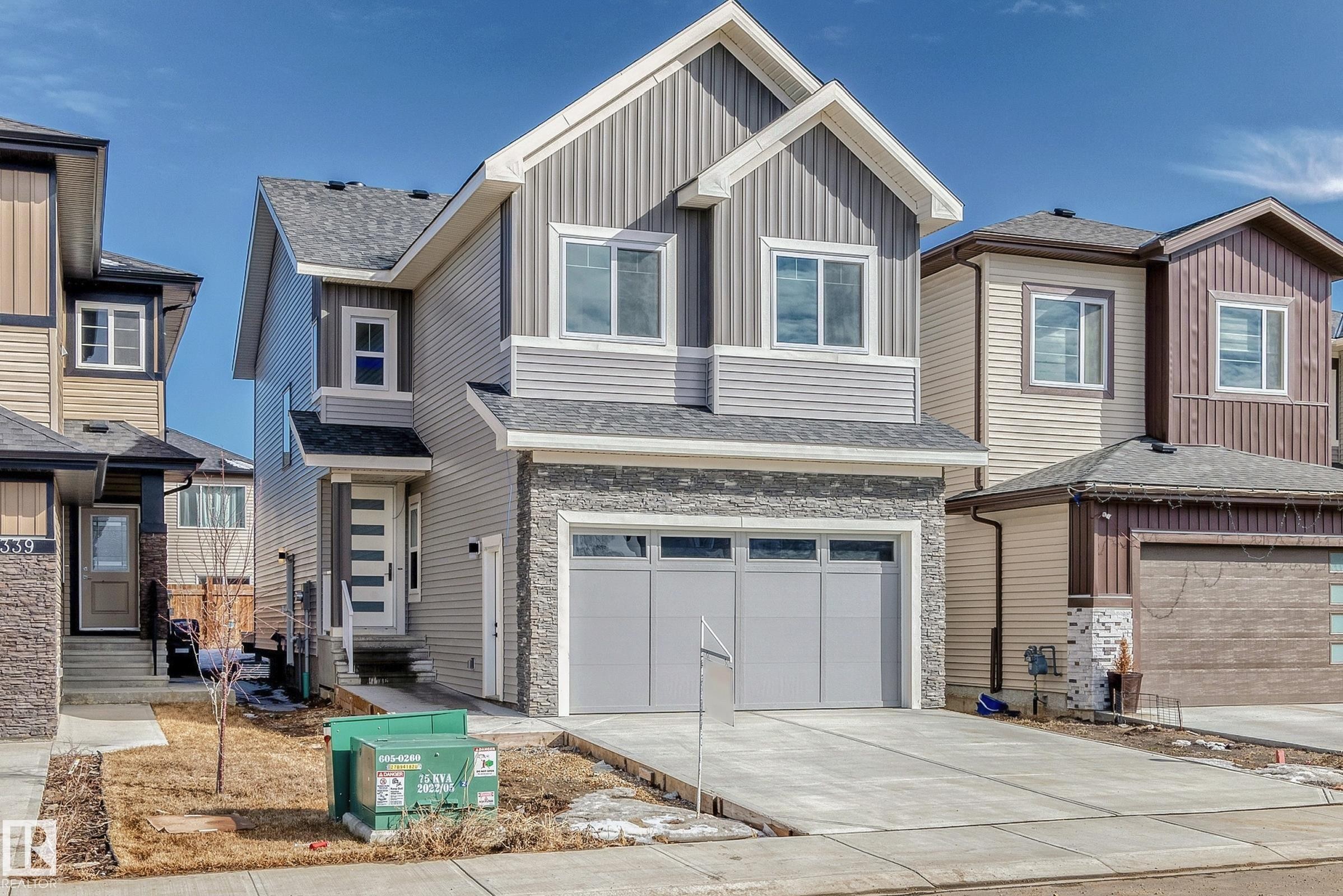 Two-story residence featuring a gray and white exterior with vertical and horizontal siding, a stone-accented garage, a contemporary frosted-glass entry door, and a concrete driveway - 341 Roberts Wynd, Leduc, AB