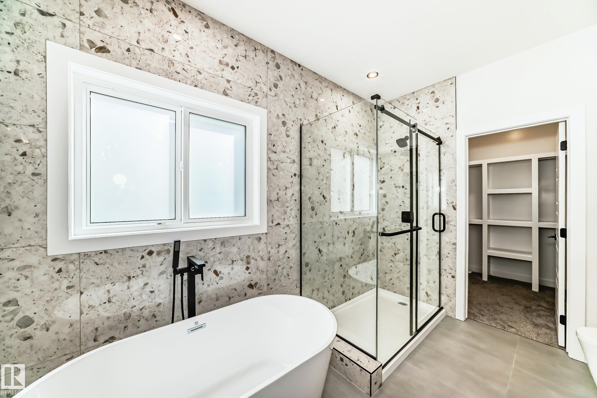Contemporary bathroom featuring a freestanding soaking tub with matte black fixtures, a walk-in shower with clear glass enclosure and black frame, large format tile flooring, and a window with white trim - 341 Roberts Wynd, Leduc, AB