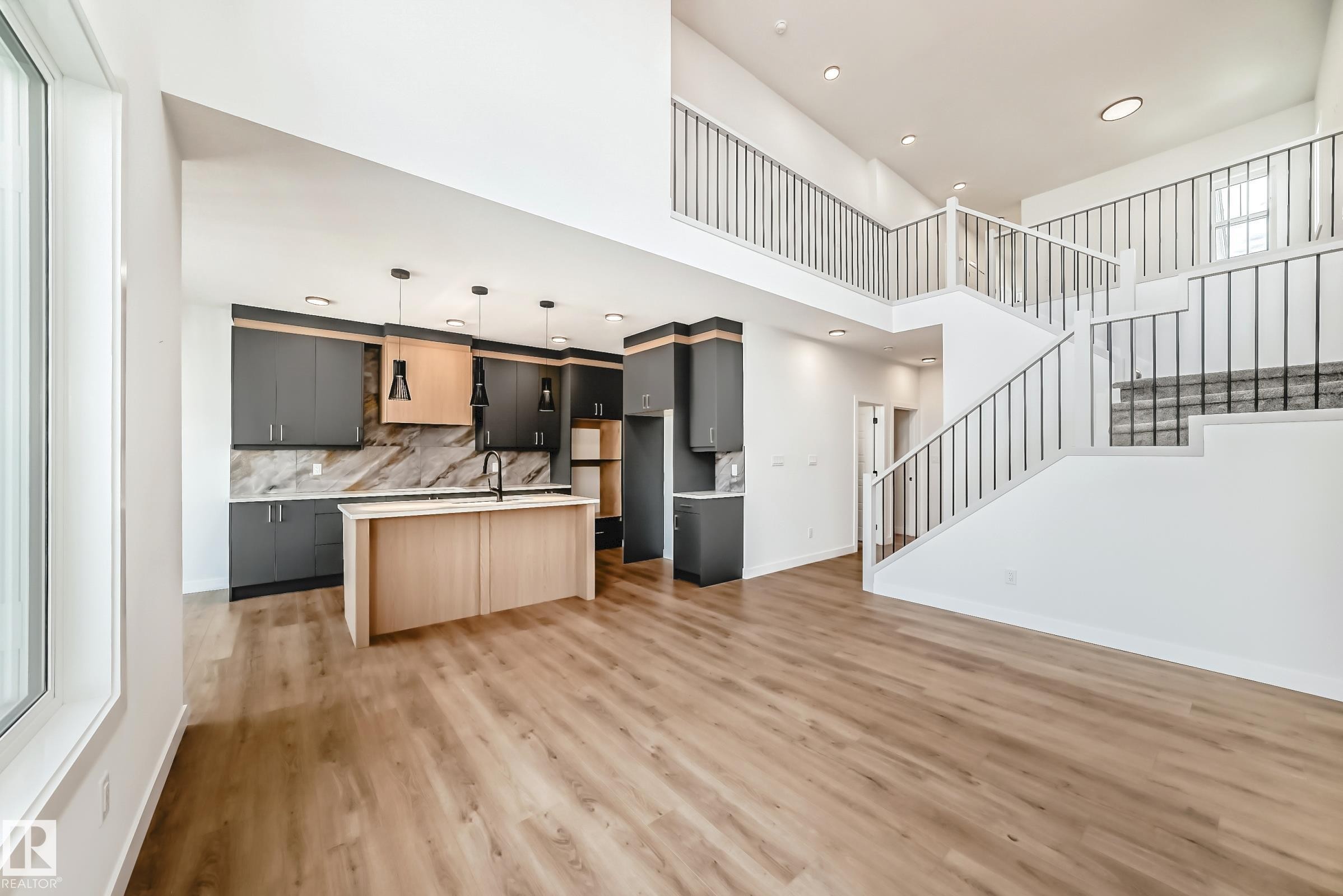 Open-concept living area featuring wood-finish flooring, a contemporary kitchen with two-tone cabinetry and a light wood-finish island, a grand staircase with metal spindles, and high ceilings - 341 Roberts Wynd, Leduc, AB