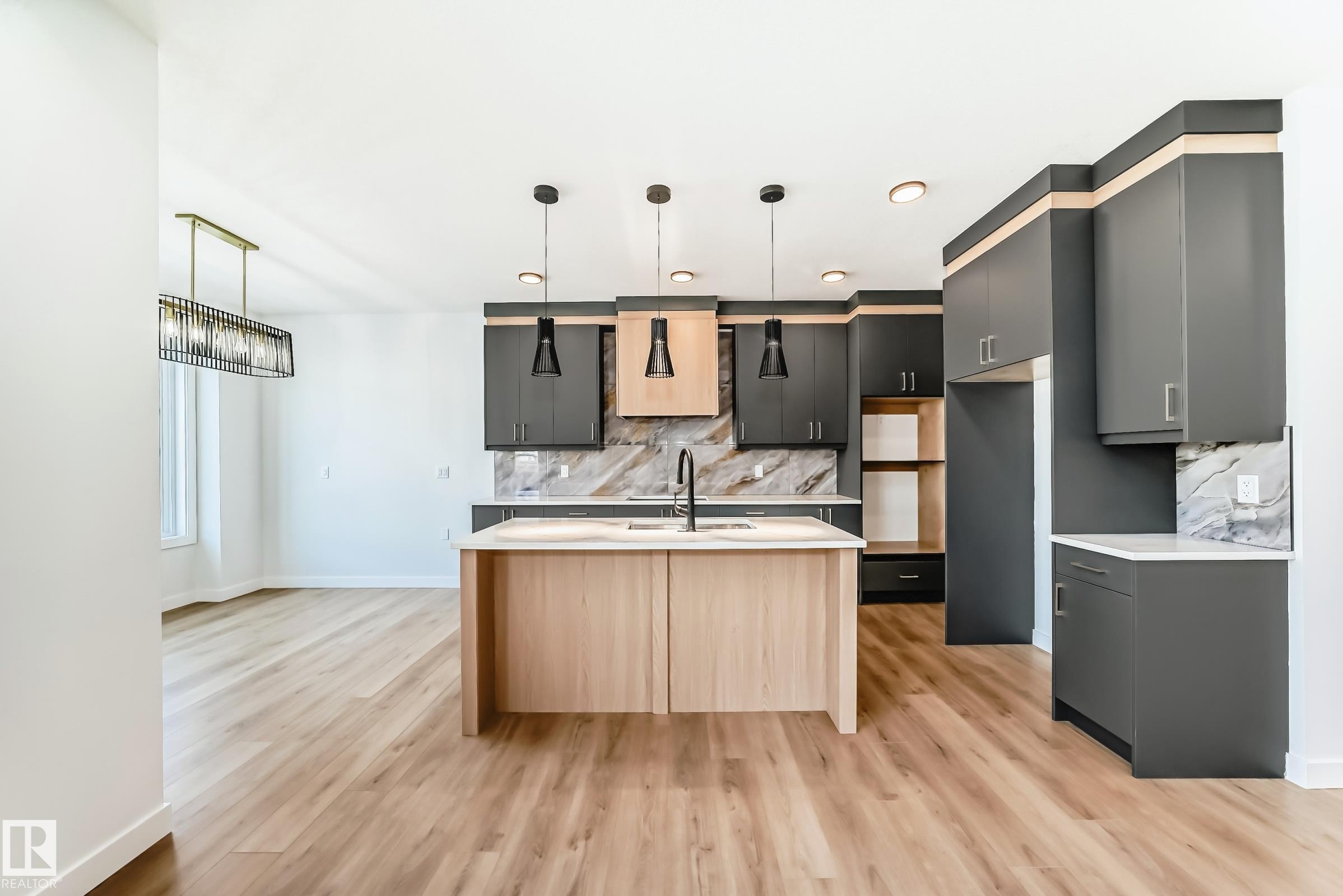 Contemporary kitchen featuring an island with integrated sink, dark cabinetry, light wood-finish accents, and a stone backsplash - 341 Roberts Wynd, Leduc, AB