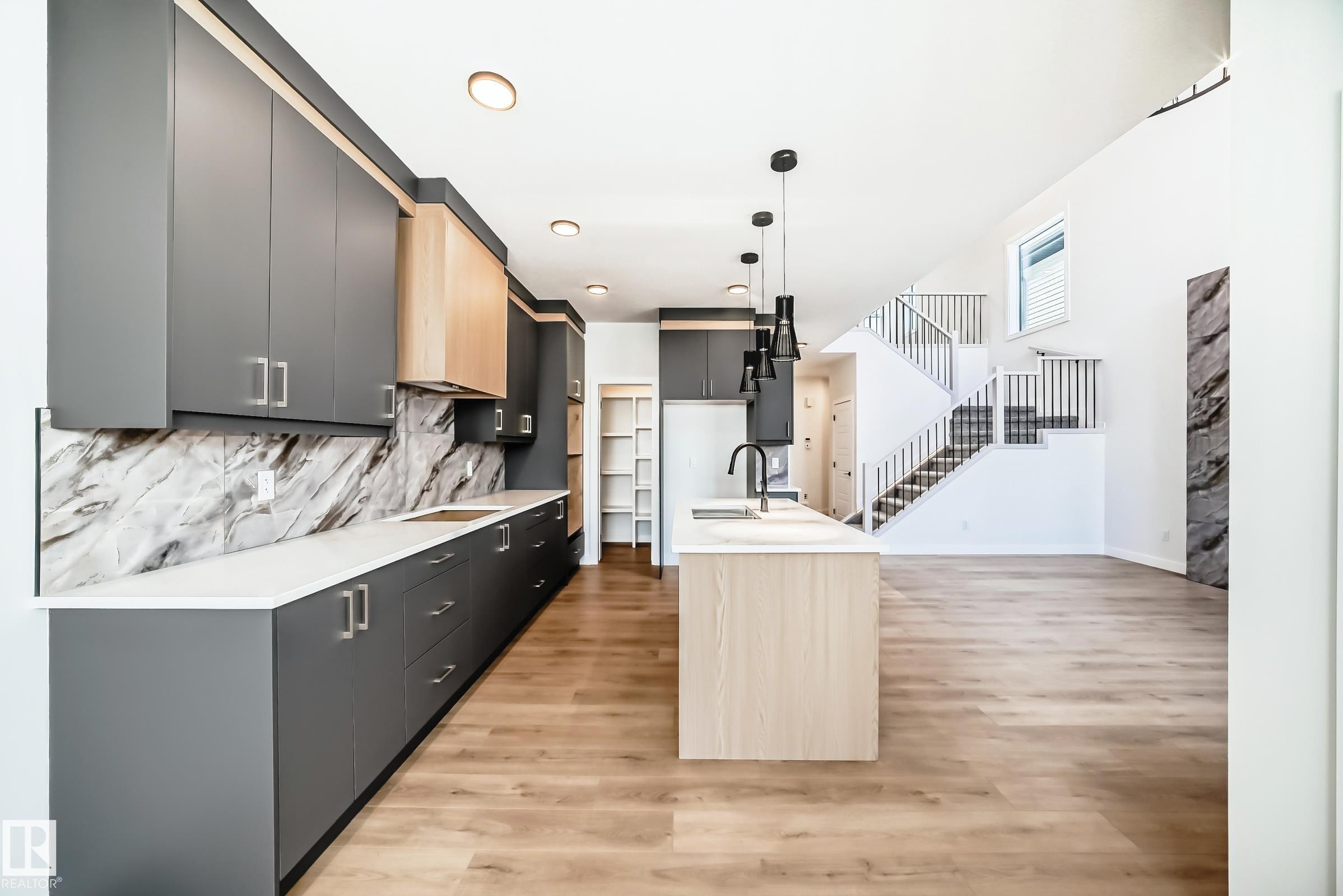 Contemporary kitchen with wood-finish flooring, an expansive island featuring a built-in sink and pendant lighting, sleek grey cabinetry, light wood-finish accents, and a distinctive marble-patterned backsplash - 341 Roberts Wynd, Leduc, AB