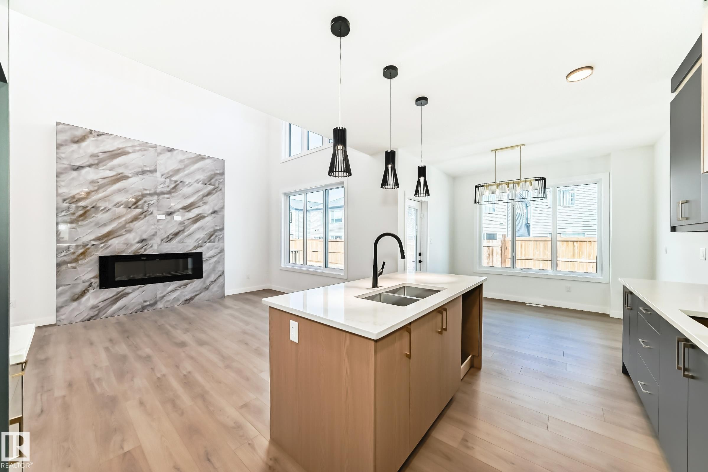 Open-concept living space featuring a central kitchen island with a double sink, a striking fireplace with a stone-finish facade, high ceilings, large windows, and wood-finish flooring throughout - 341 Roberts Wynd, Leduc, AB