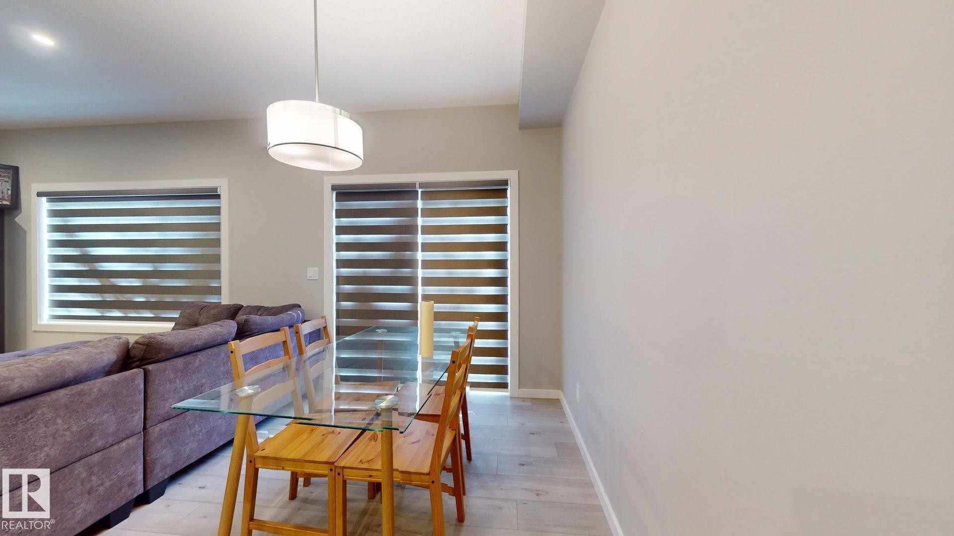 Open-concept living space featuring light wood-finish flooring, a contemporary drum pendant light, and a patio door with horizontal blinds - 607 31 Avenue, Edmonton, AB