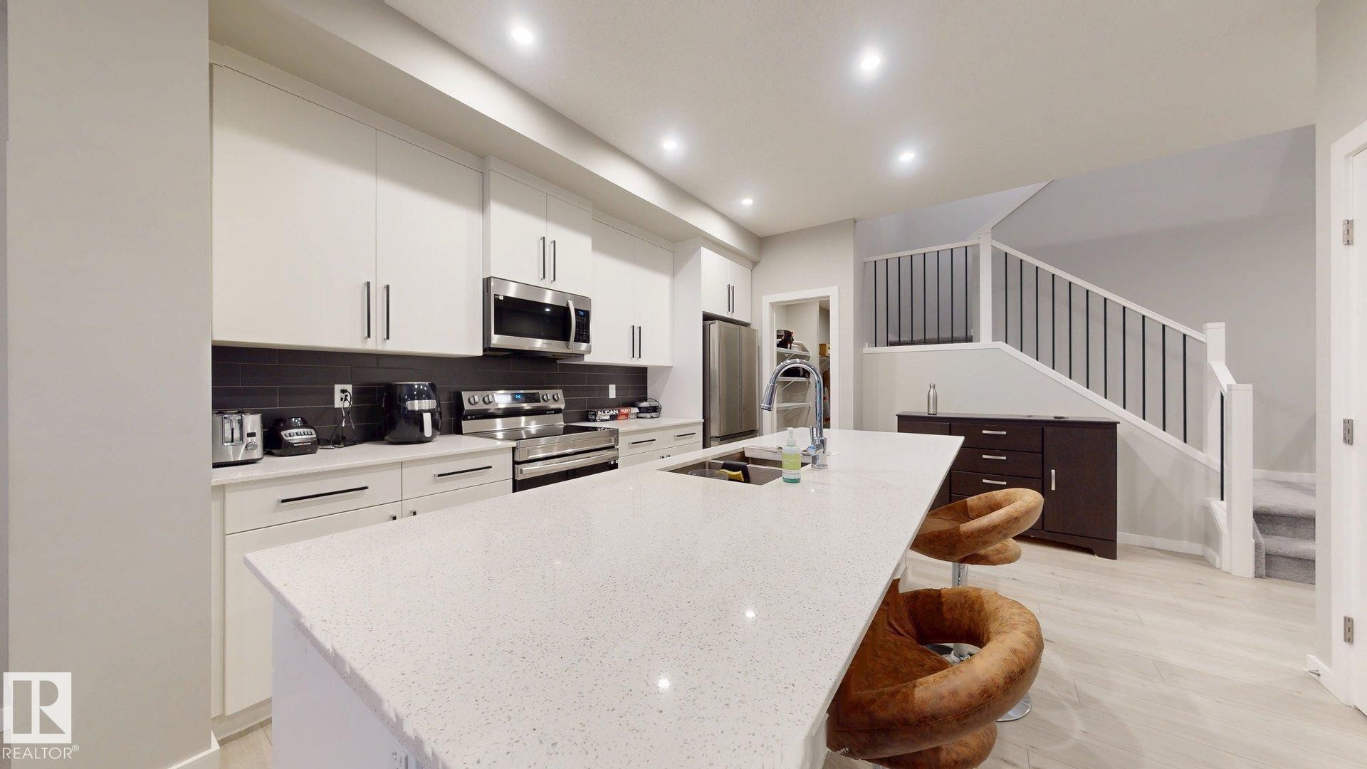 Modern kitchen featuring a large island with an undermount sink, white cabinetry with dark hardware, stainless steel appliances, dark subway tile backsplash, and recessed lighting - 607 31 Avenue, Edmonton, AB