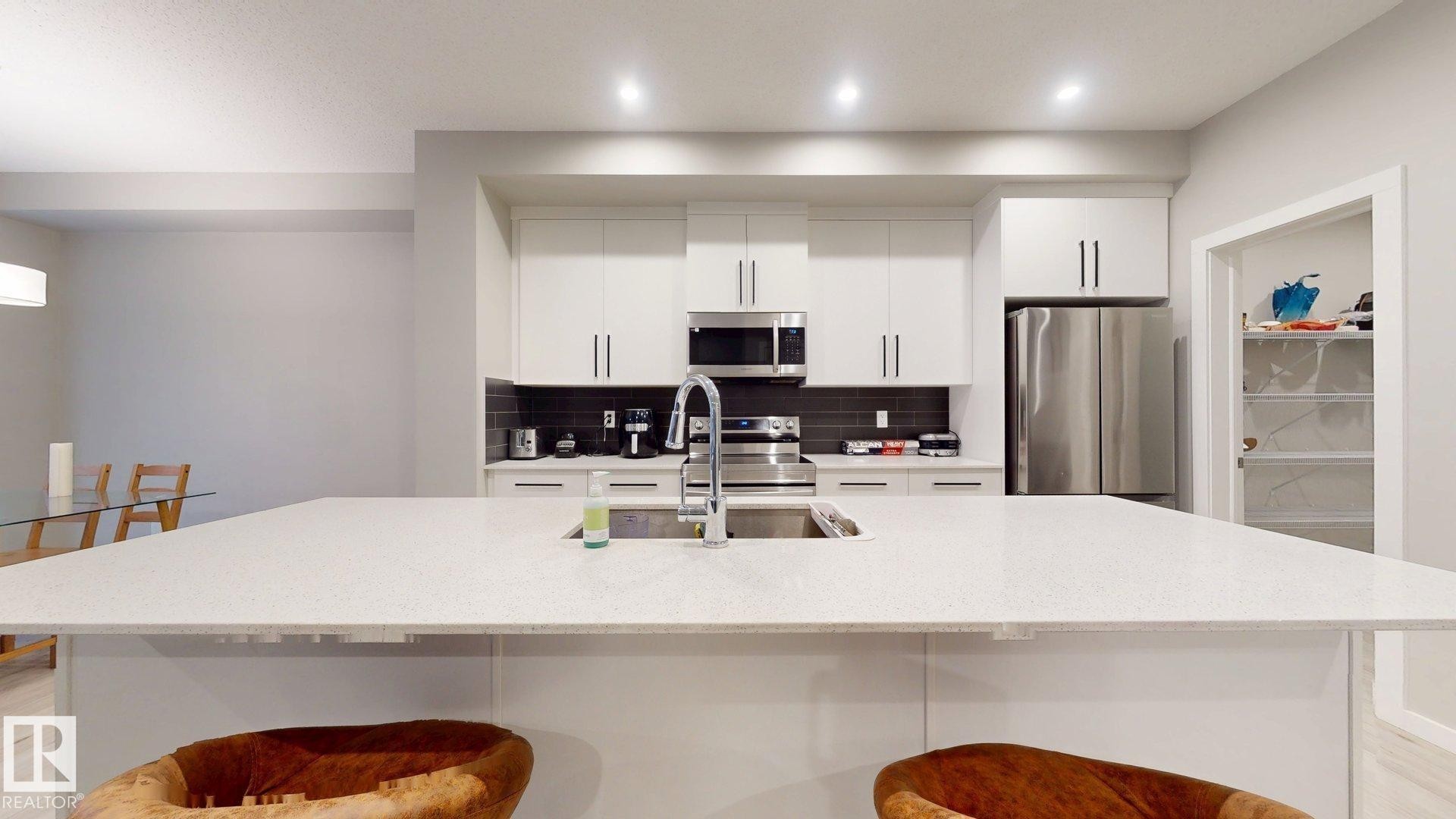 Kitchen featuring a large central island with an integrated sink and faucet, white cabinetry with dark hardware, stainless steel appliances, and a dark tile backsplash - 607 31 Avenue, Edmonton, AB