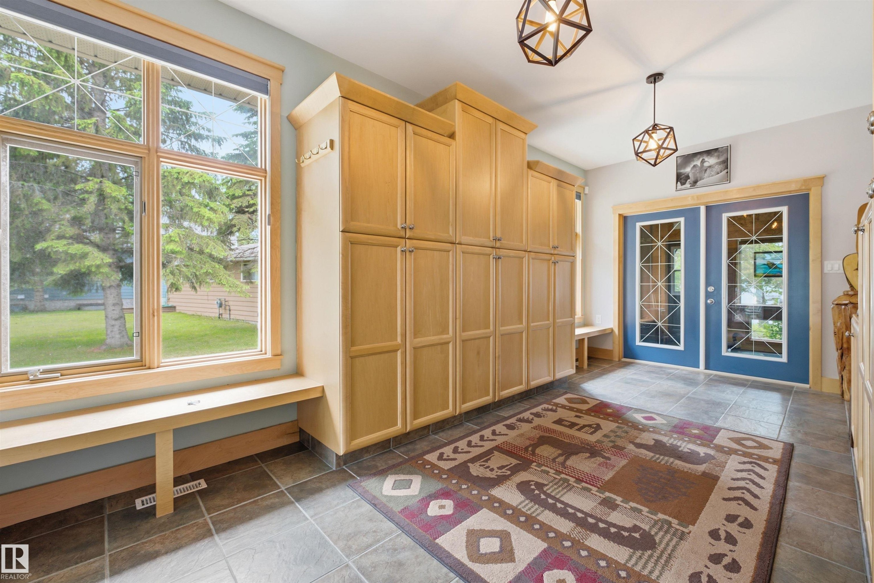 Entryway featuring extensive wood-finish cabinetry, a large window with a built-in bench, two geometric pendant lights, and blue double doors with decorative glass inserts - A38 Bernice Ave, Rural Leduc County, AB