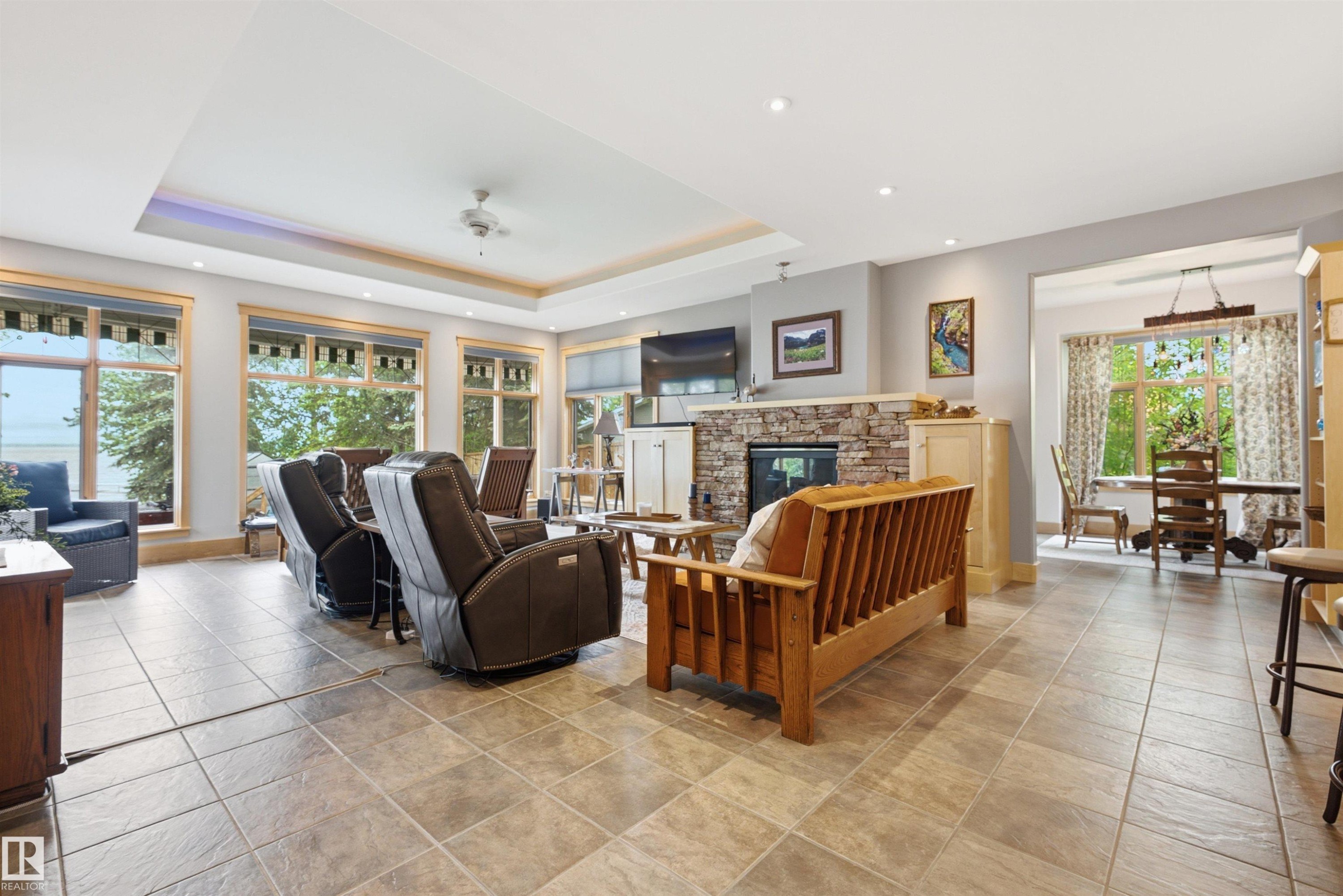 Spacious great room featuring large windows, a tray ceiling with recessed lighting, a stone-front fireplace, and large format tile flooring - A38 Bernice Ave, Rural Leduc County, AB
