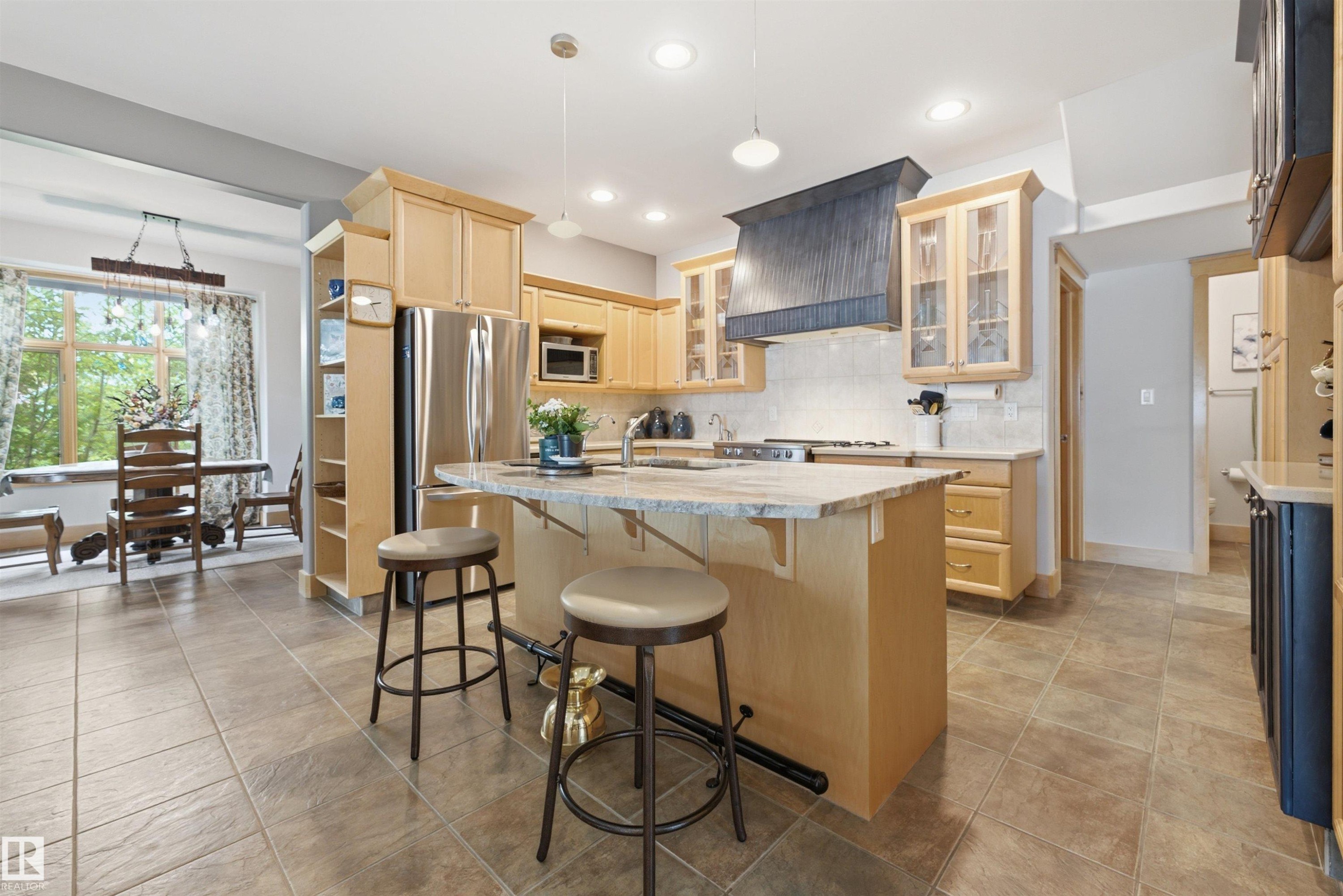 Open-concept kitchen featuring light wood cabinetry, a central island with a stone countertop, stainless steel appliances, and tile flooring - A38 Bernice Ave, Rural Leduc County, AB