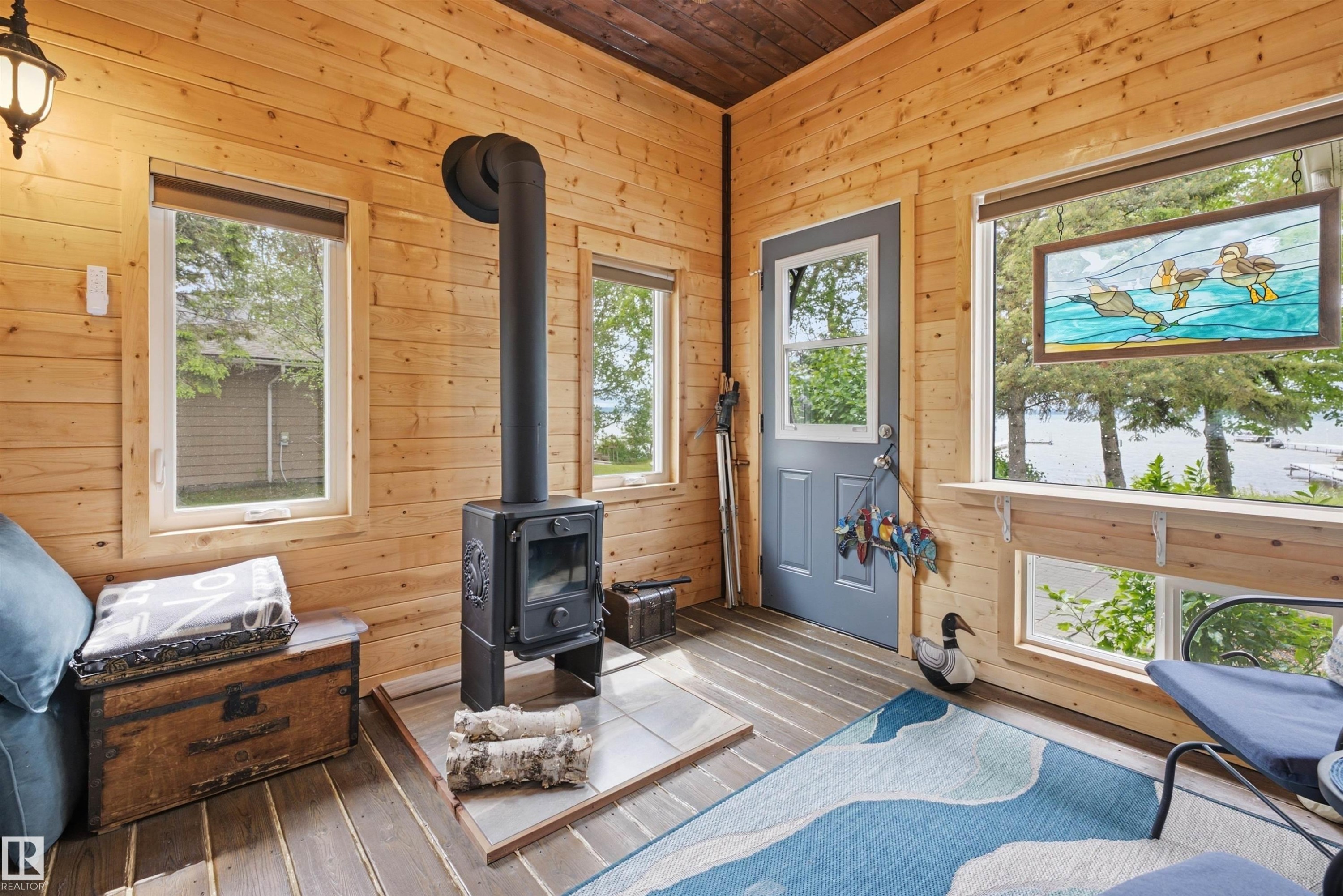 Inviting interior space featuring wood-paneled walls and ceiling, a black freestanding wood-burning stove on a tiled hearth, multiple windows, and a light blue exterior door - A38 Bernice Ave, Rural Leduc County, AB