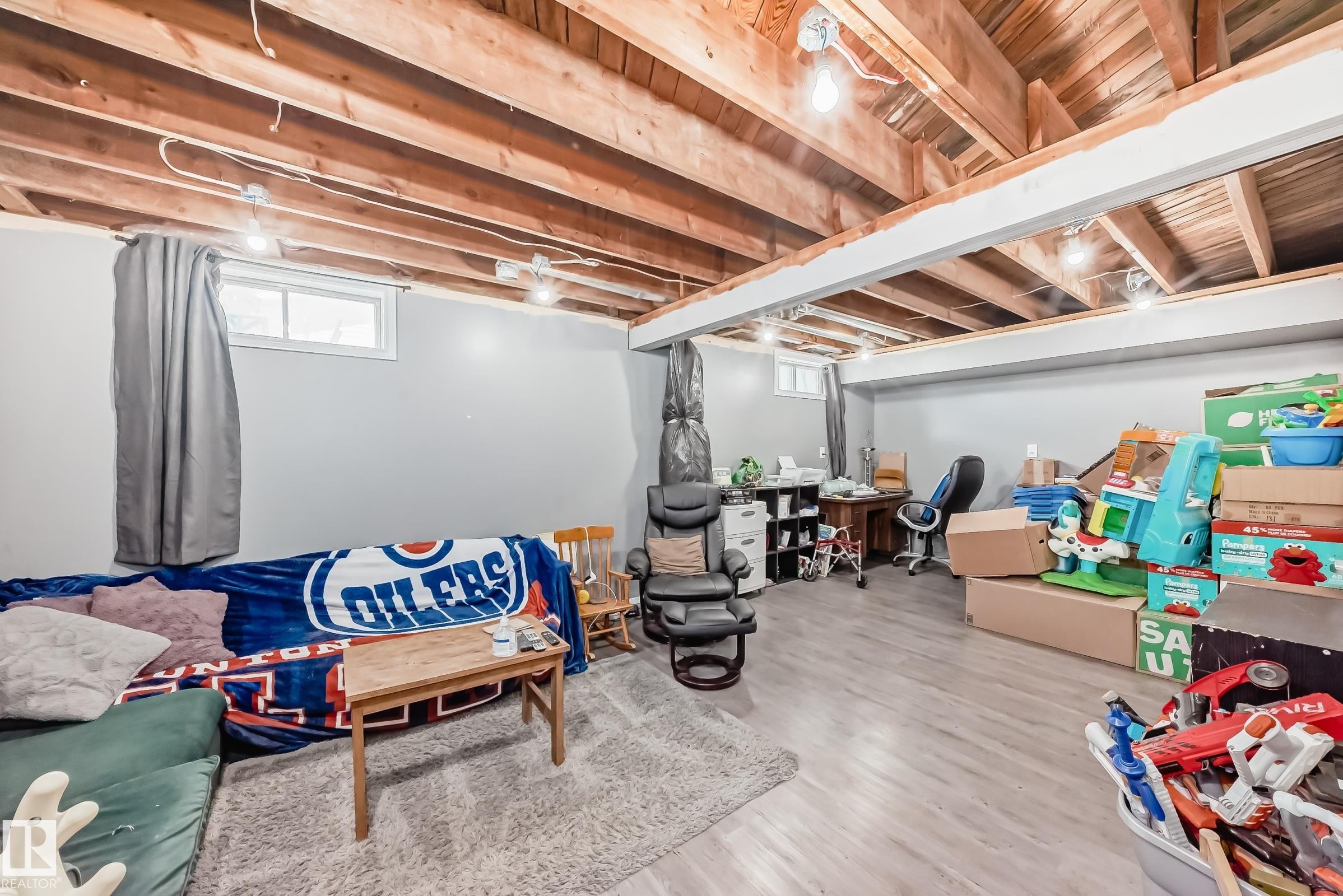 Spacious lower-level room featuring light gray walls, exposed wood ceiling joists, and light wood-finish flooring - 5106 19 Avenue, Edmonton, AB