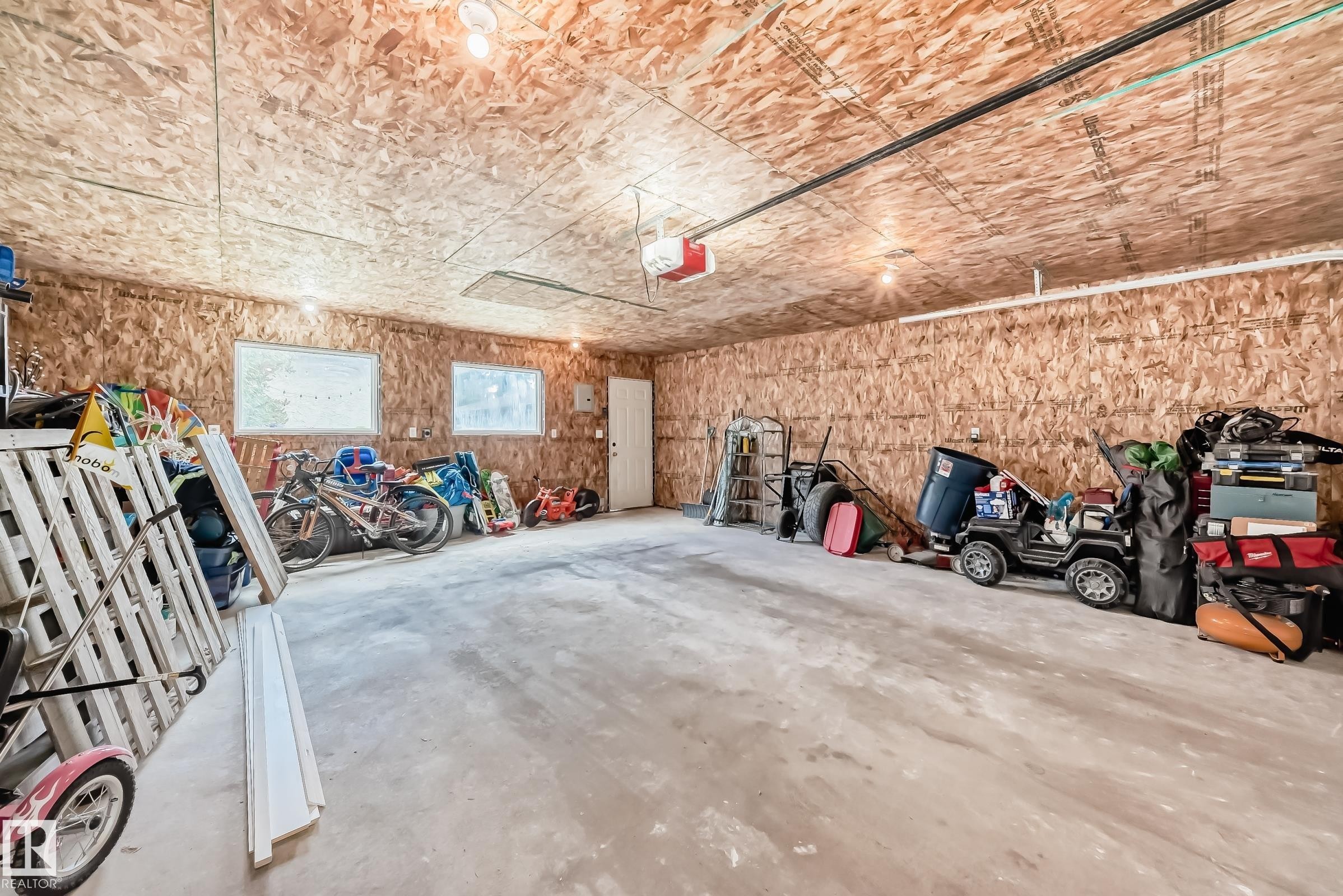 Spacious garage featuring a concrete floor, exposed OSB walls and ceiling, multiple windows, overhead lighting, and an overhead garage door opener mechanism - 5106 19 Avenue, Edmonton, AB