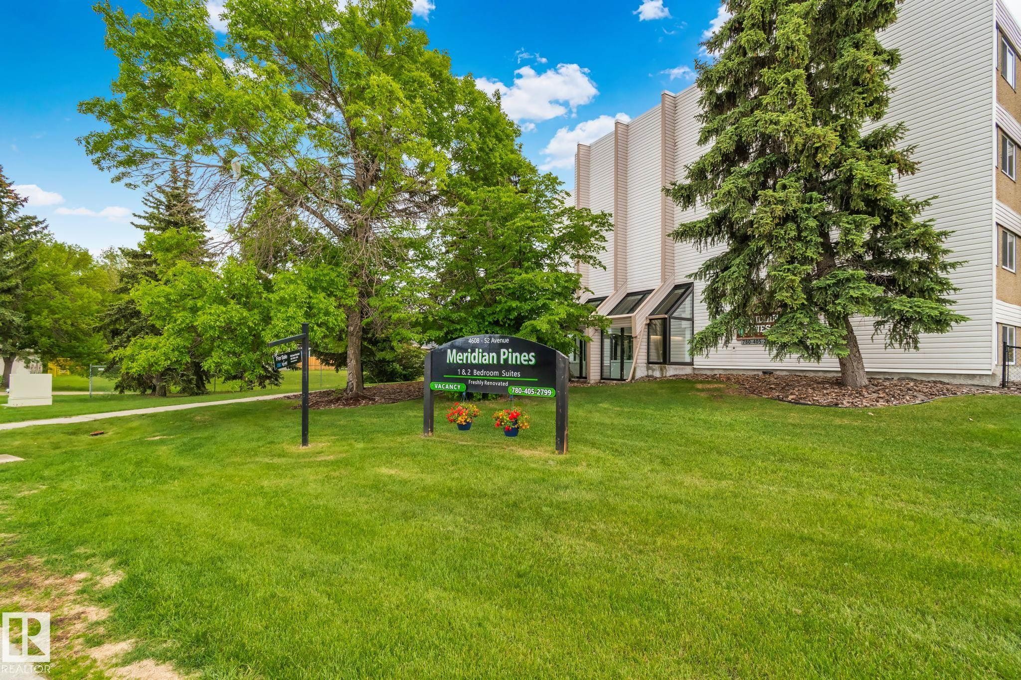 Multi-story building featuring a white and tan exterior, a glass-enclosed entrance, and mature trees on a manicured lawn - 115 4608 52 Avenue, Stony Plain, AB