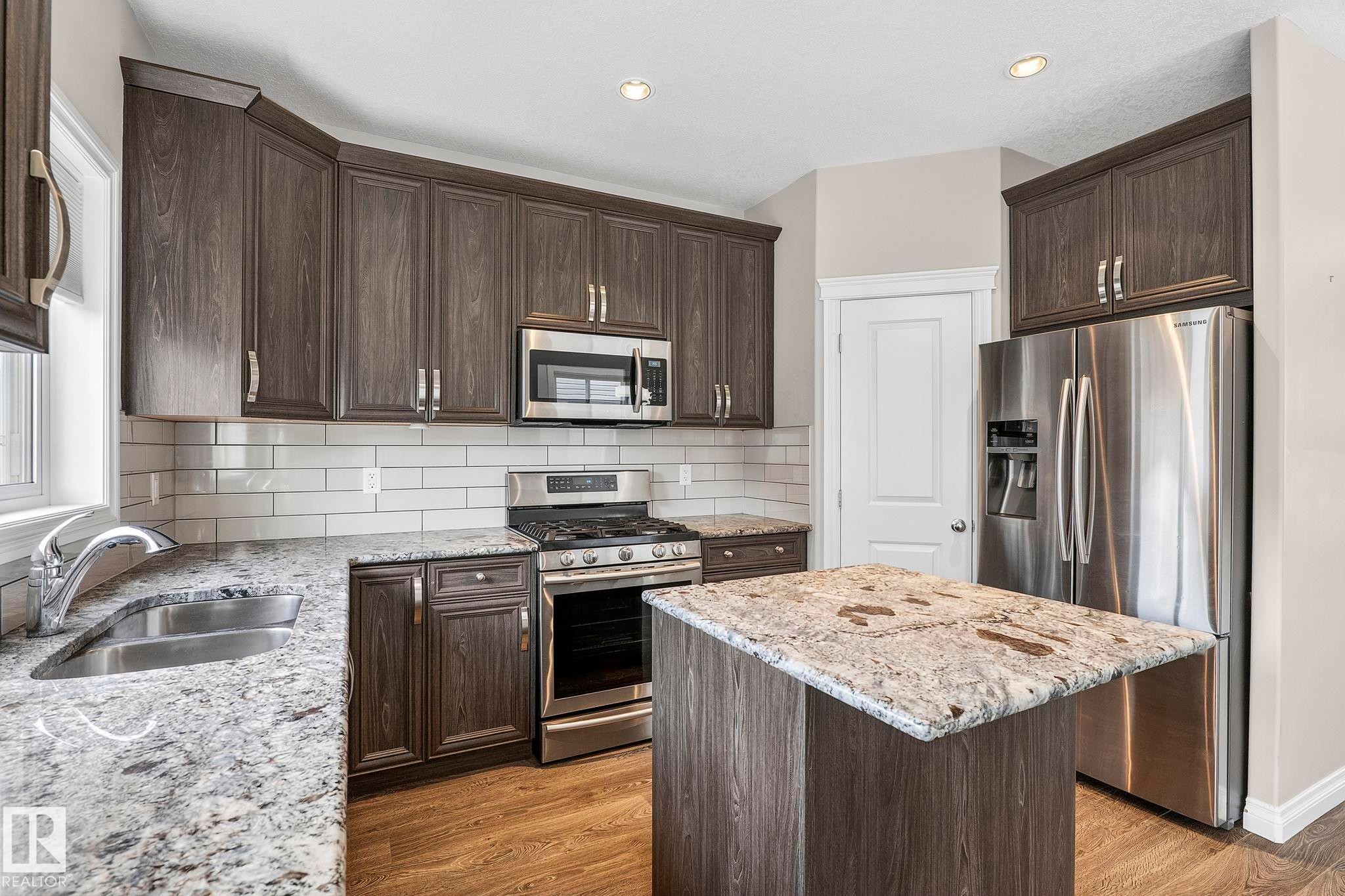 Modern kitchen featuring dark wood-finish cabinetry, stainless steel appliances, speckled countertops, a white subway tile backsplash, and wood-finish flooring - 6927 Cardinal Wynd Sw, Edmonton, AB - Indoor Photo Showing Kitchen With Double Sink