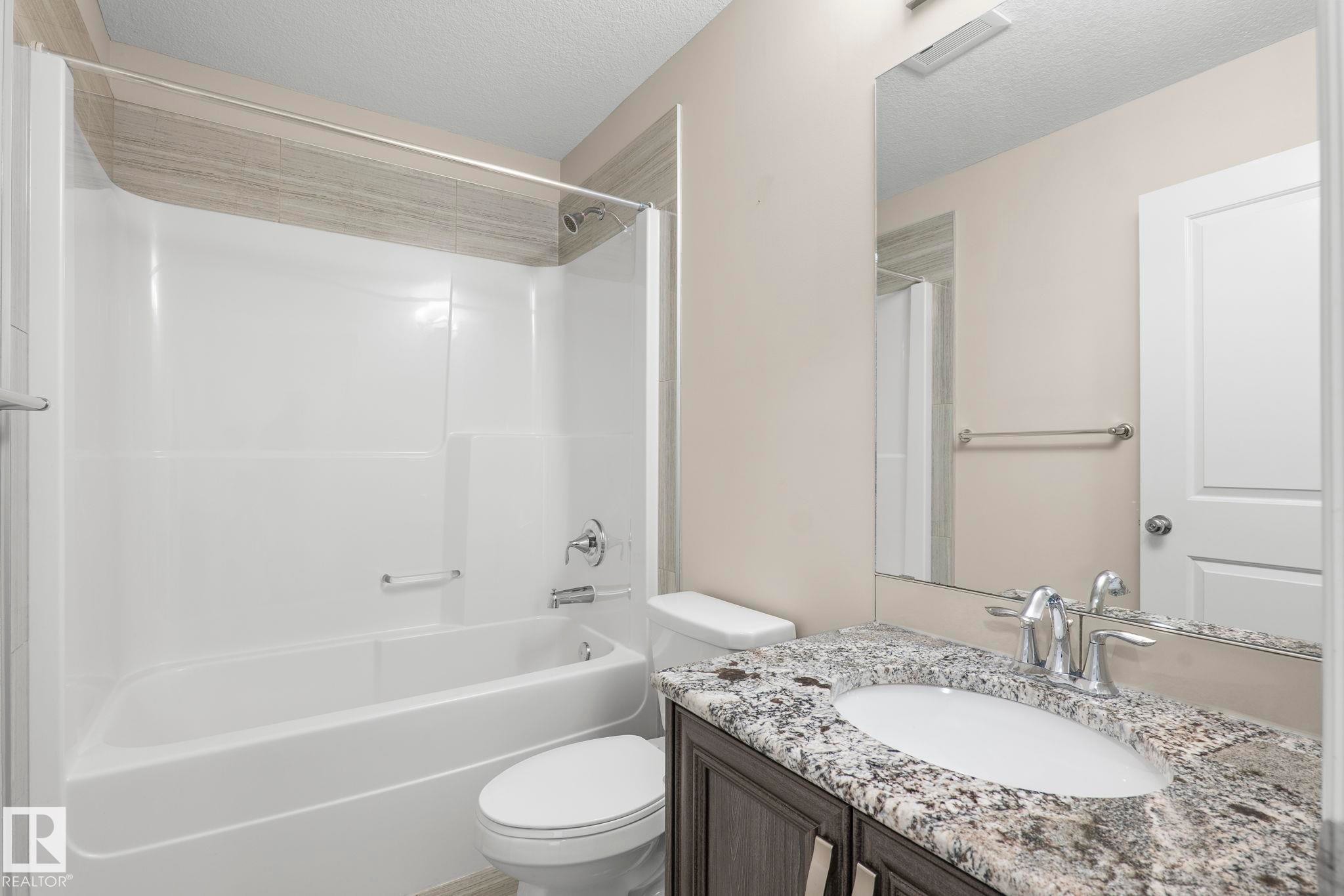 Bathroom featuring a white tub/shower combination, decorative tile surround, dark wood-finish vanity with an undermount sink, and a speckled countertop - 6927 Cardinal Wynd Sw, Edmonton, AB - Indoor Photo Showing Bathroom