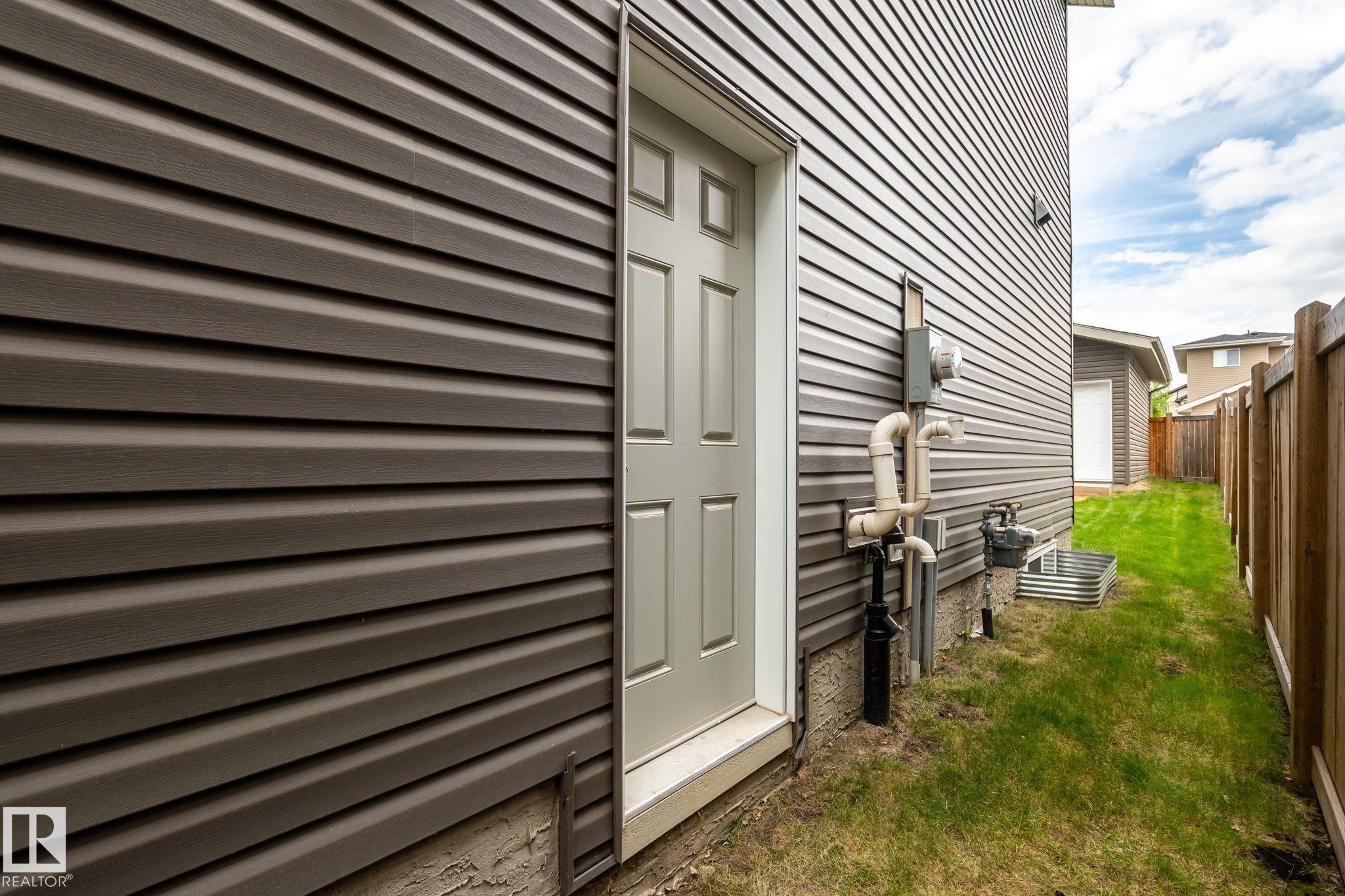 Exterior side entrance with a paneled door, dark horizontal siding, established grass, and a wooden fence - 6927 Cardinal Wynd Sw, Edmonton, AB - Outdoor With Exterior