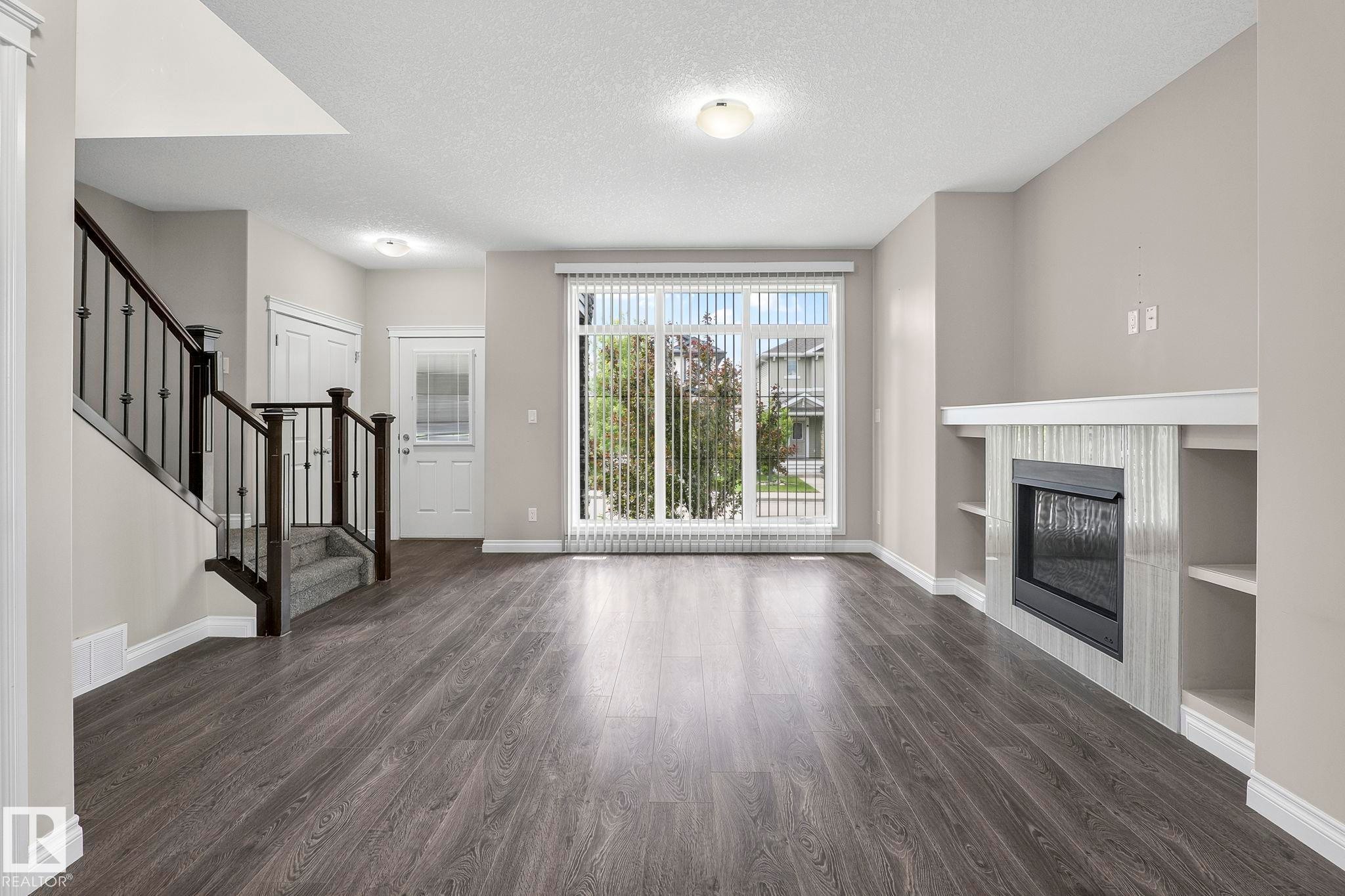 Spacious living area featuring wood-finish flooring, a contemporary fireplace with a tiled surround, built-in shelving, and a large window with vertical blinds - 6927 Cardinal Wynd Sw, Edmonton, AB - Indoor With Fireplace