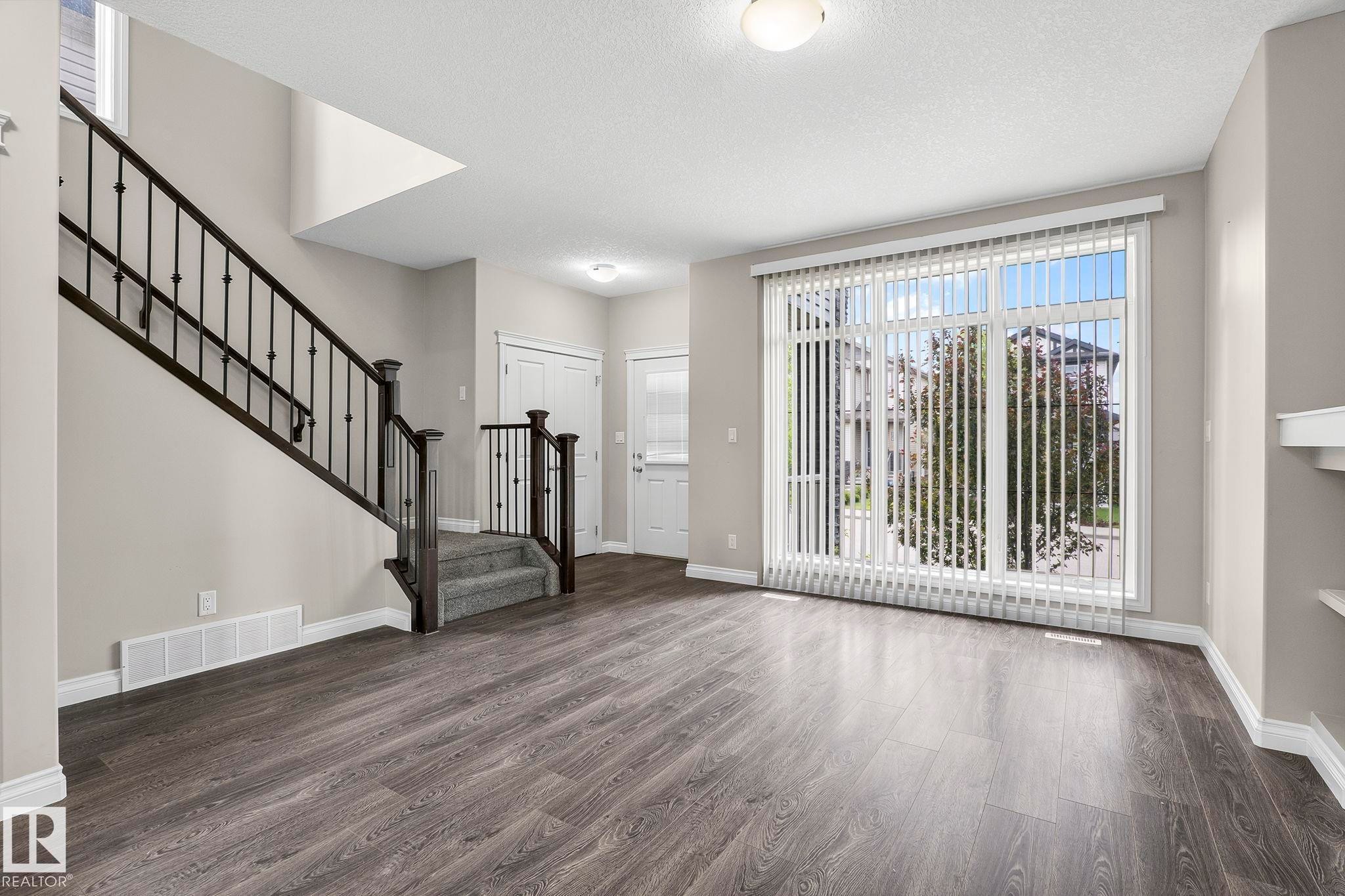 Spacious living area featuring wood-finish flooring, a contemporary staircase with wrought iron spindles, a large window with vertical blinds, and neutral wall paint - 6927 Cardinal Wynd Sw, Edmonton, AB - Indoor Photo Showing Other Room