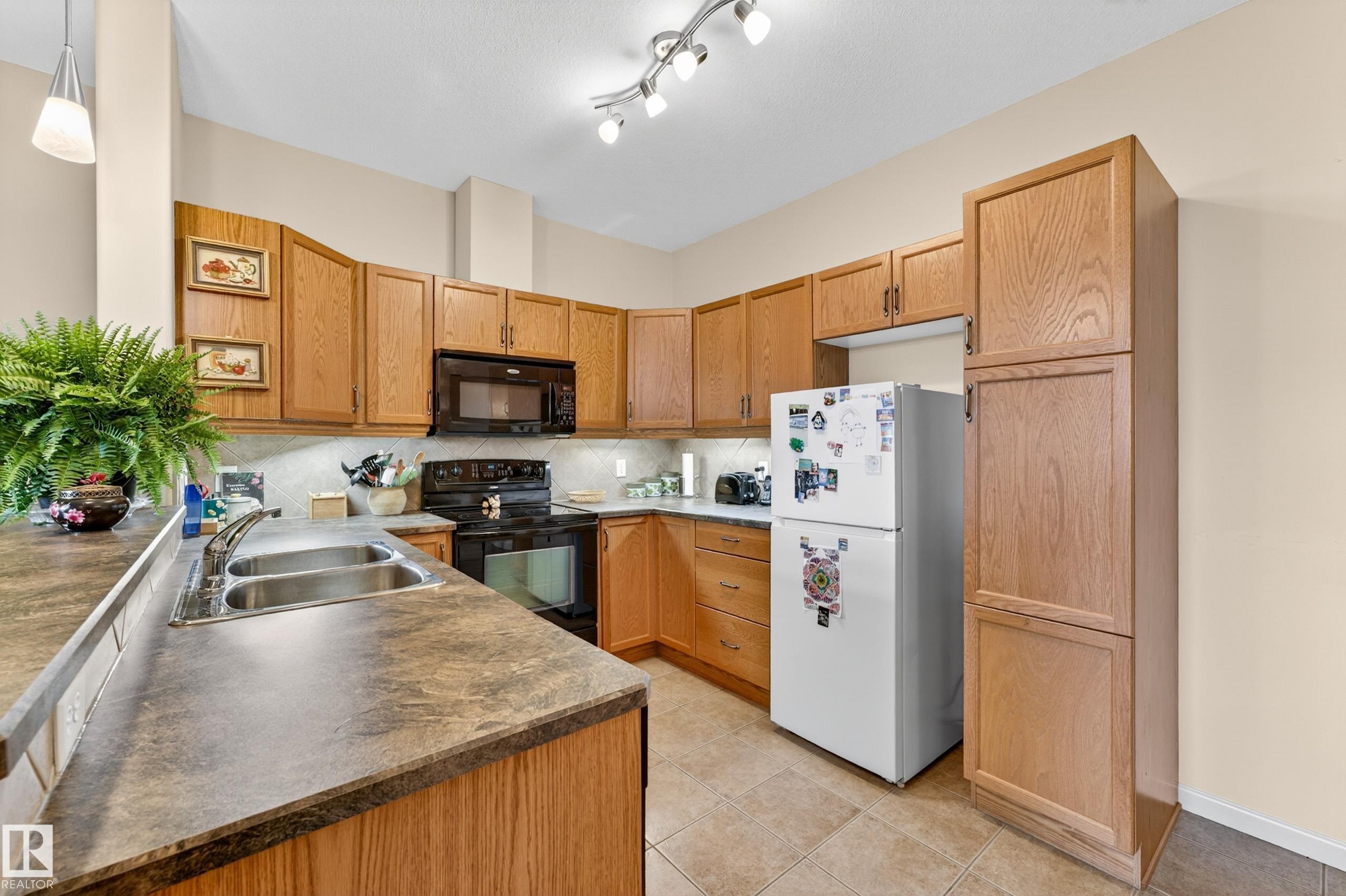 Kitchen featuring wood cabinetry, a double basin stainless steel sink, black appliances, and tiled flooring - 133 2503 Hanna Crescent, Edmonton, AB