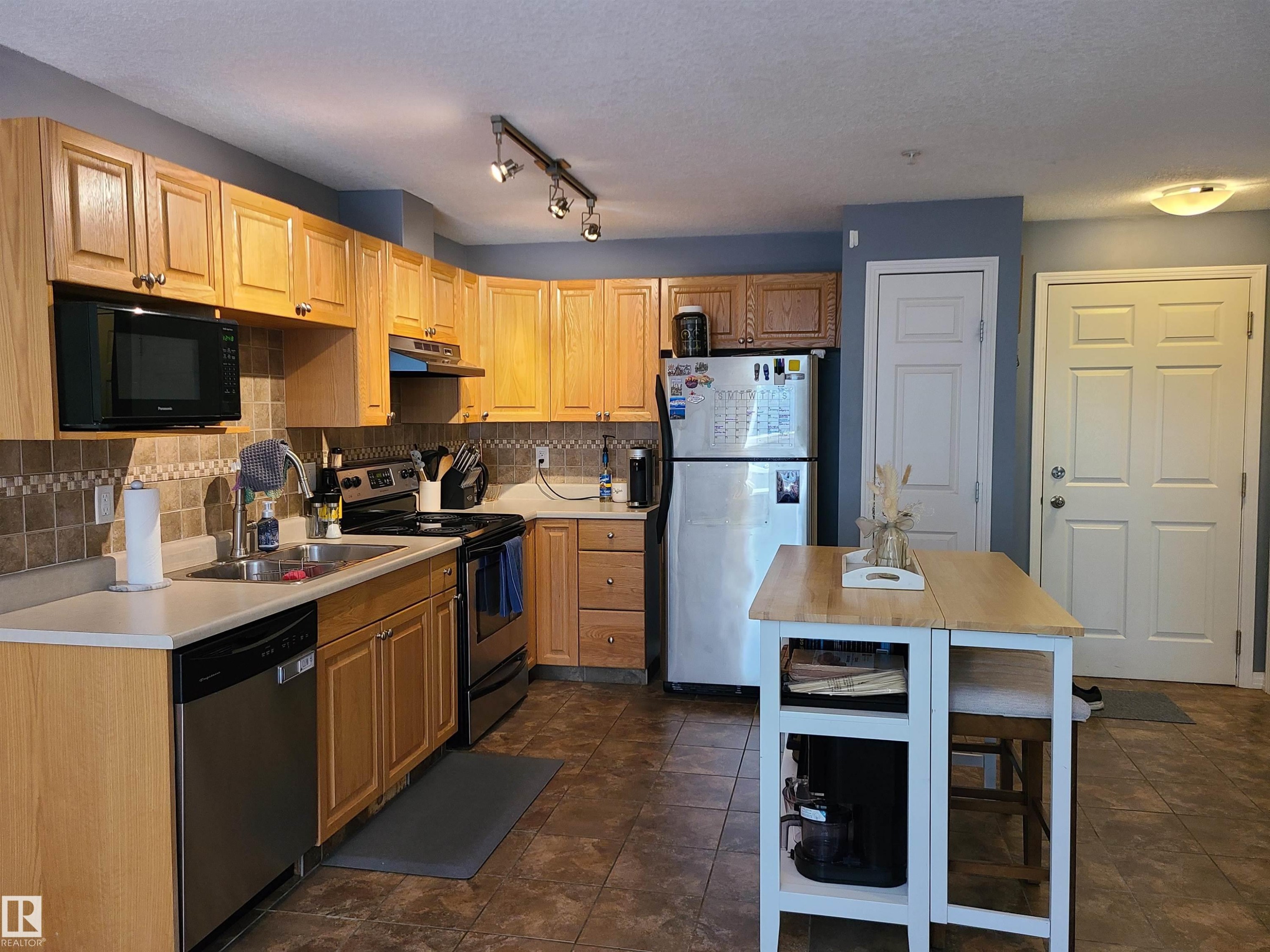 Kitchen featuring wood cabinetry, stainless steel appliances, a tiled backsplash, and stone-finish flooring - 116 5005 165 Avenue, Edmonton, AB