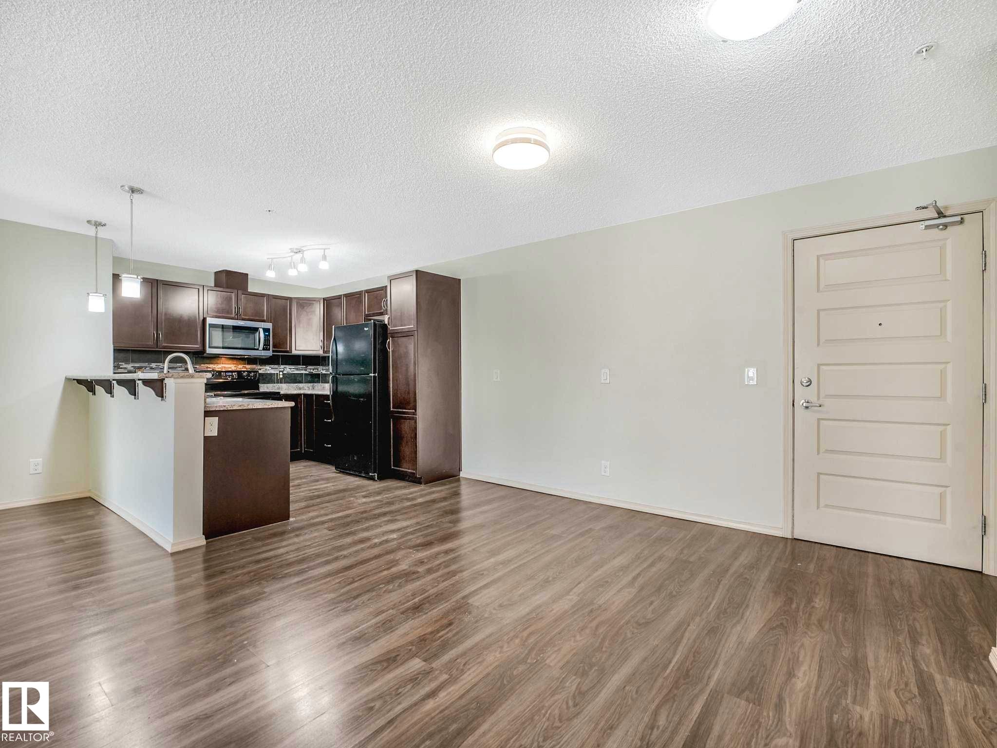 Open-concept living area featuring wood-finish flooring, a kitchen with dark cabinetry, black appliances, and pendant lighting - 117 6070 Schonsee Way, Edmonton, AB