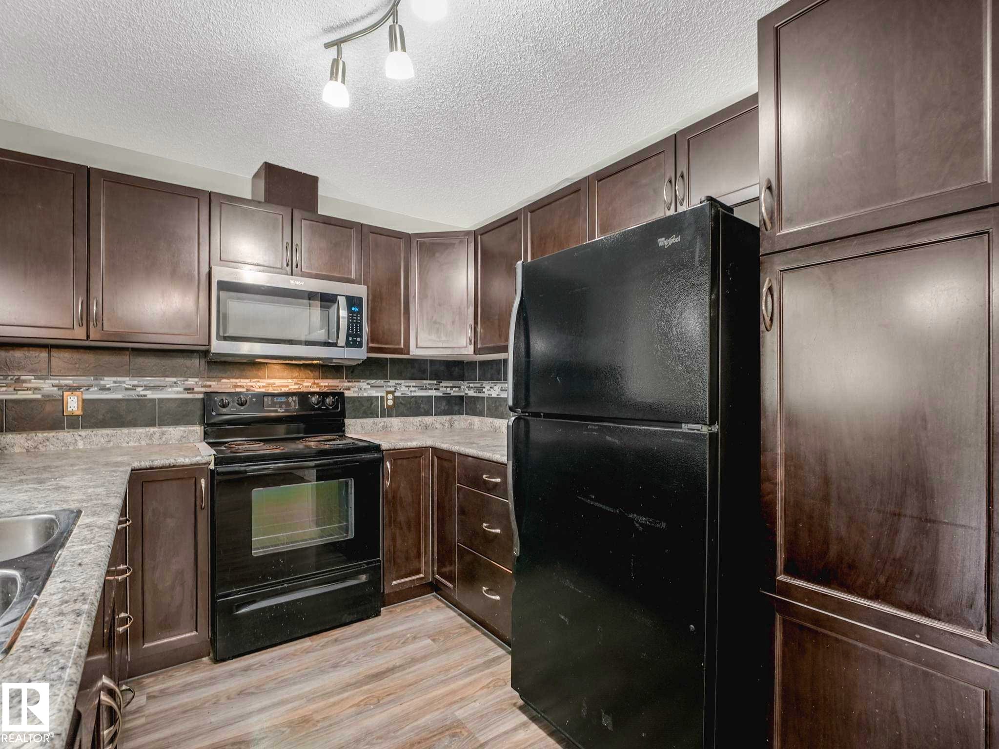 Kitchen featuring dark wood cabinetry, a stainless steel microwave, black electric range, a black refrigerator, and wood-finish flooring - 117 6070 Schonsee Way, Edmonton, AB
