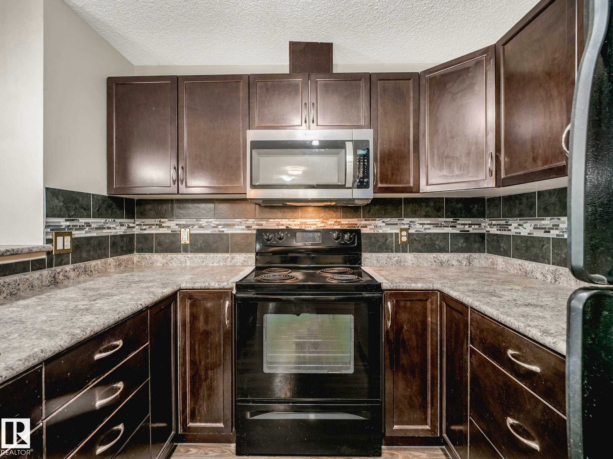 U-shaped kitchen featuring dark wood cabinetry, a stainless steel microwave, black electric range, and a decorative tile backsplash with mosaic accents - 117 6070 Schonsee Way, Edmonton, AB