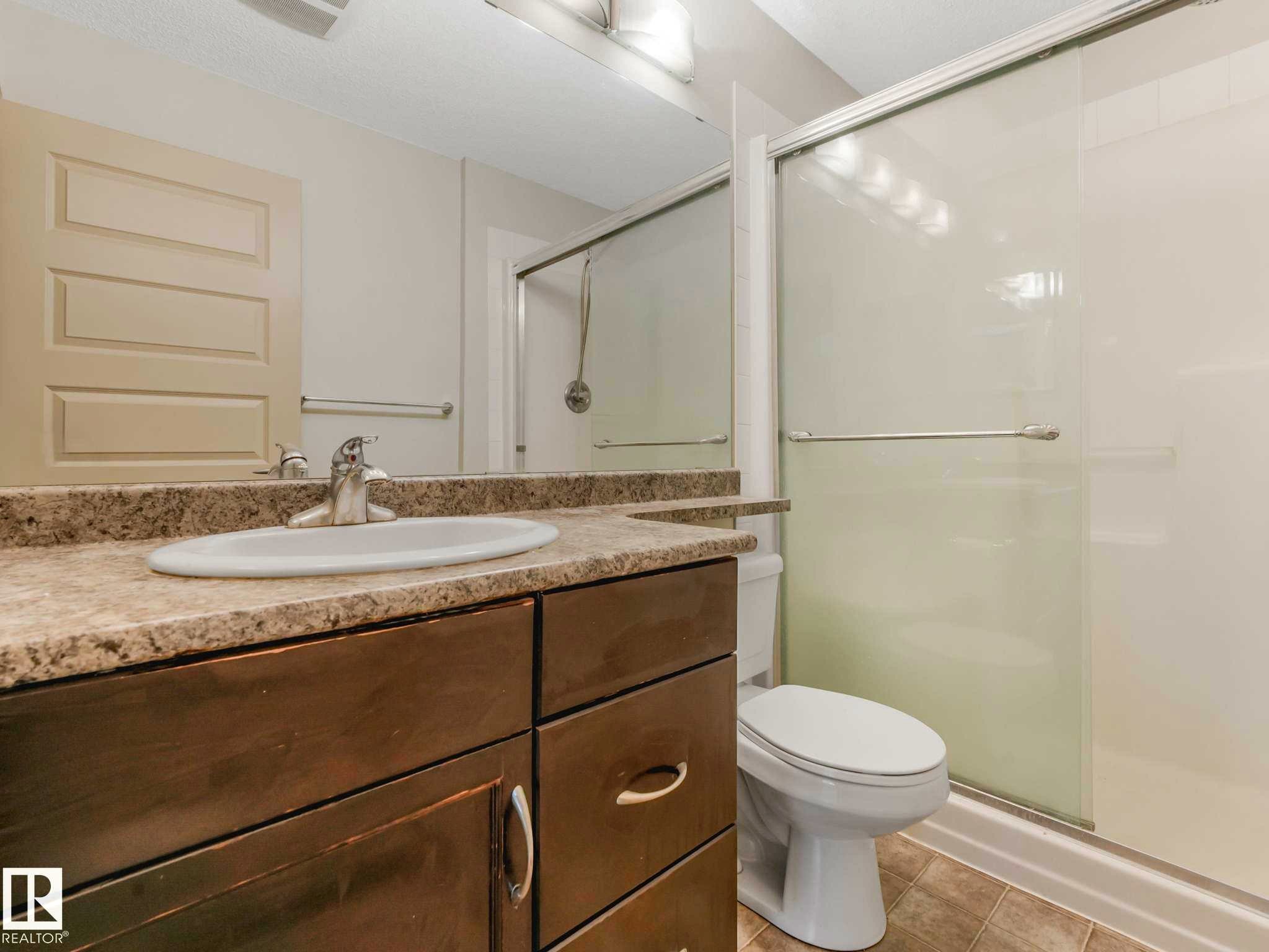 Bathroom featuring a dark wood-finish vanity with an integrated sink, a neutral-toned countertop, a shower with a sliding glass door, and tiled flooring - 117 6070 Schonsee Way, Edmonton, AB