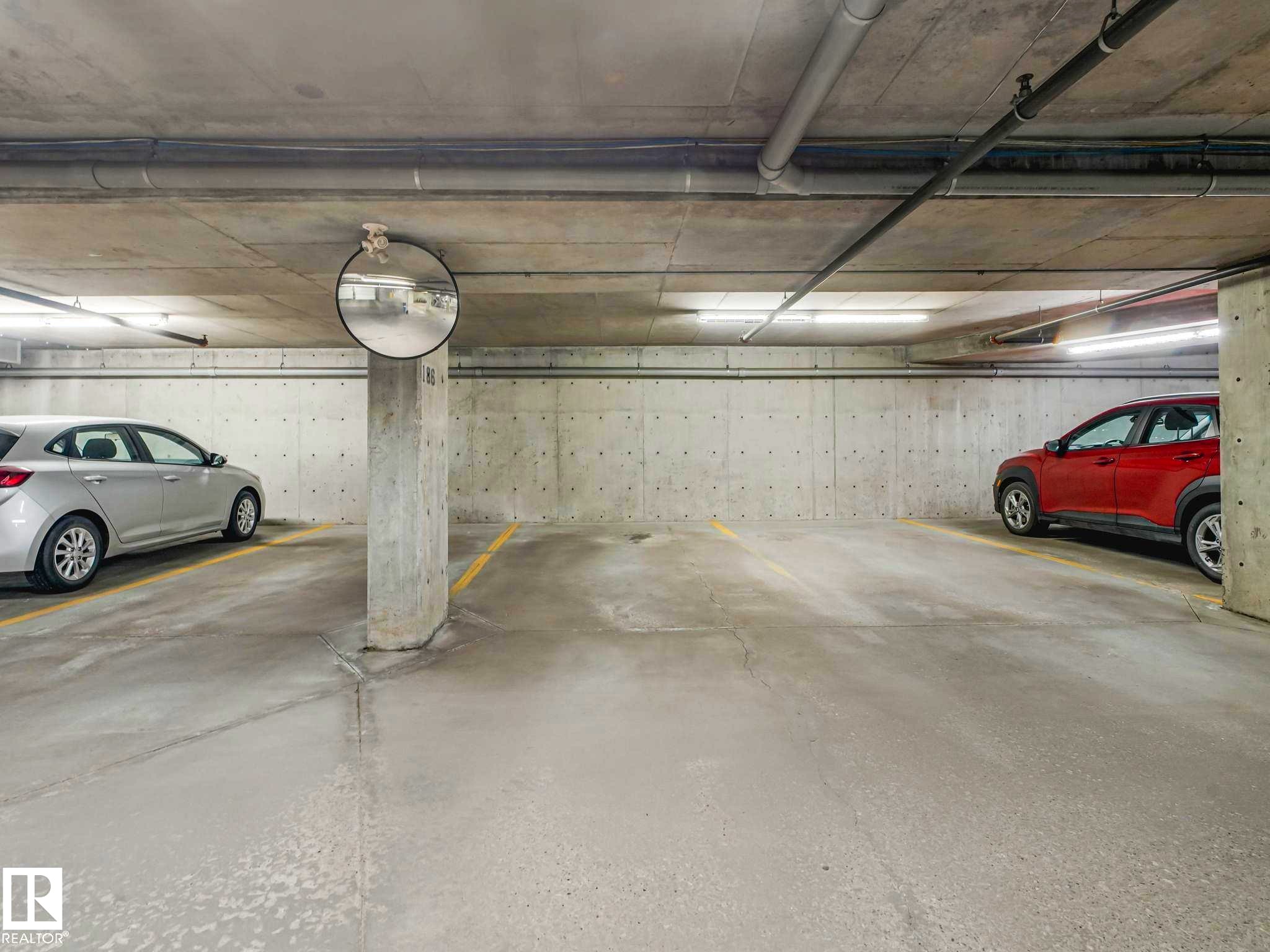 Underground parking garage featuring concrete walls and ceiling, painted concrete flooring, overhead fluorescent lighting, and a convex mirror for visibility - 117 6070 Schonsee Way, Edmonton, AB