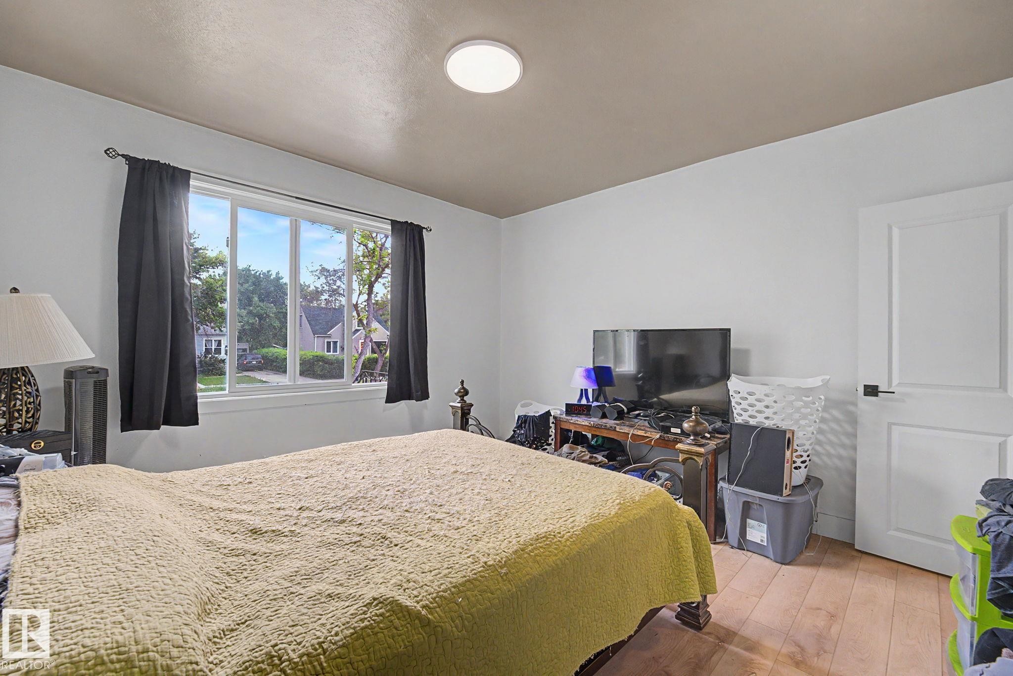 Spacious room featuring a large window, a modern ceiling light fixture, wood-finish flooring, and a paneled interior door with black hardware - 11914 124 Street, Edmonton, AB
