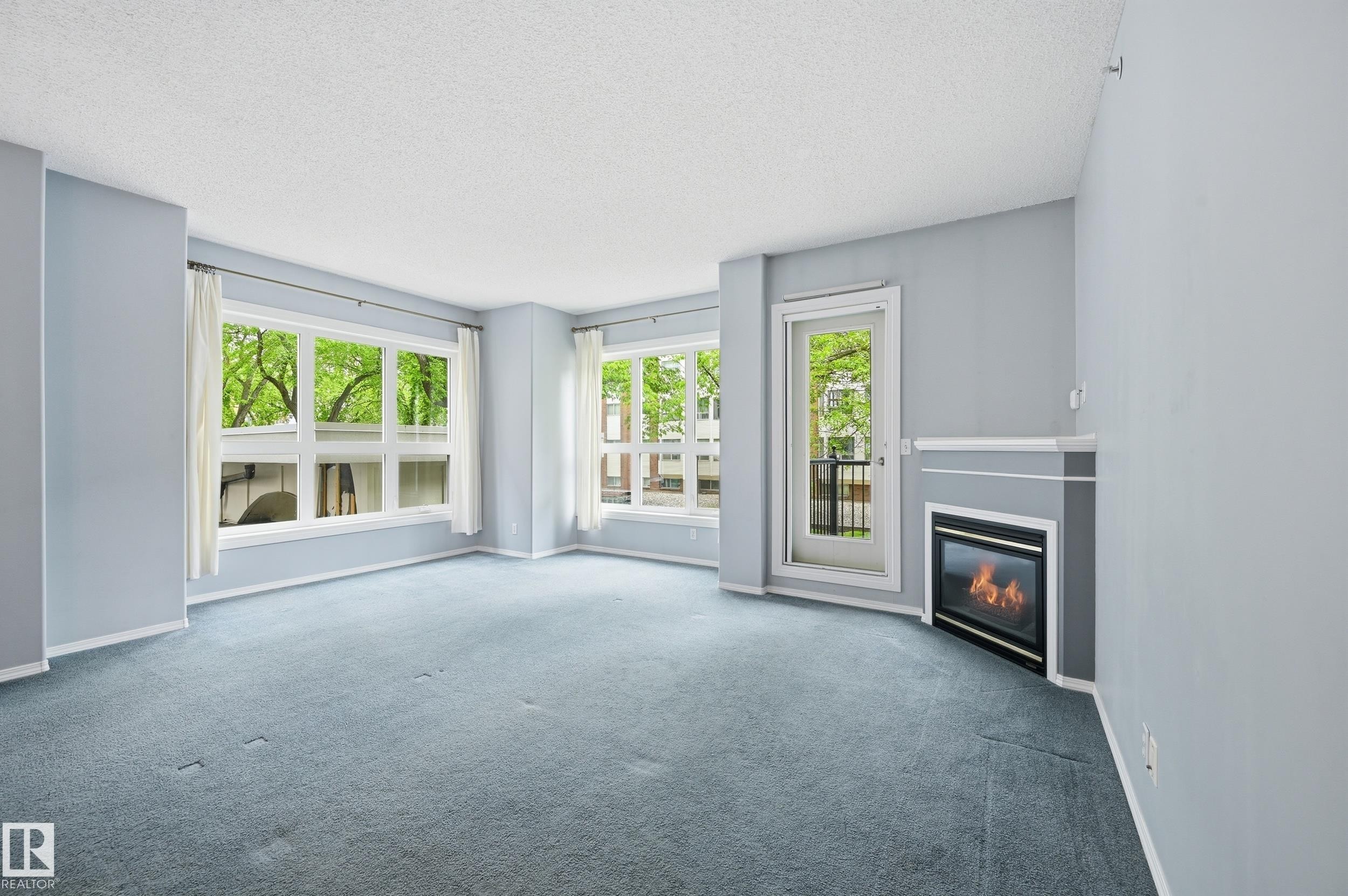 Living area featuring an integrated fireplace with a corner mantel, light blue carpeting, multiple windows, and a glass-paneled door - 203 11103 84 Avenue, Edmonton, AB